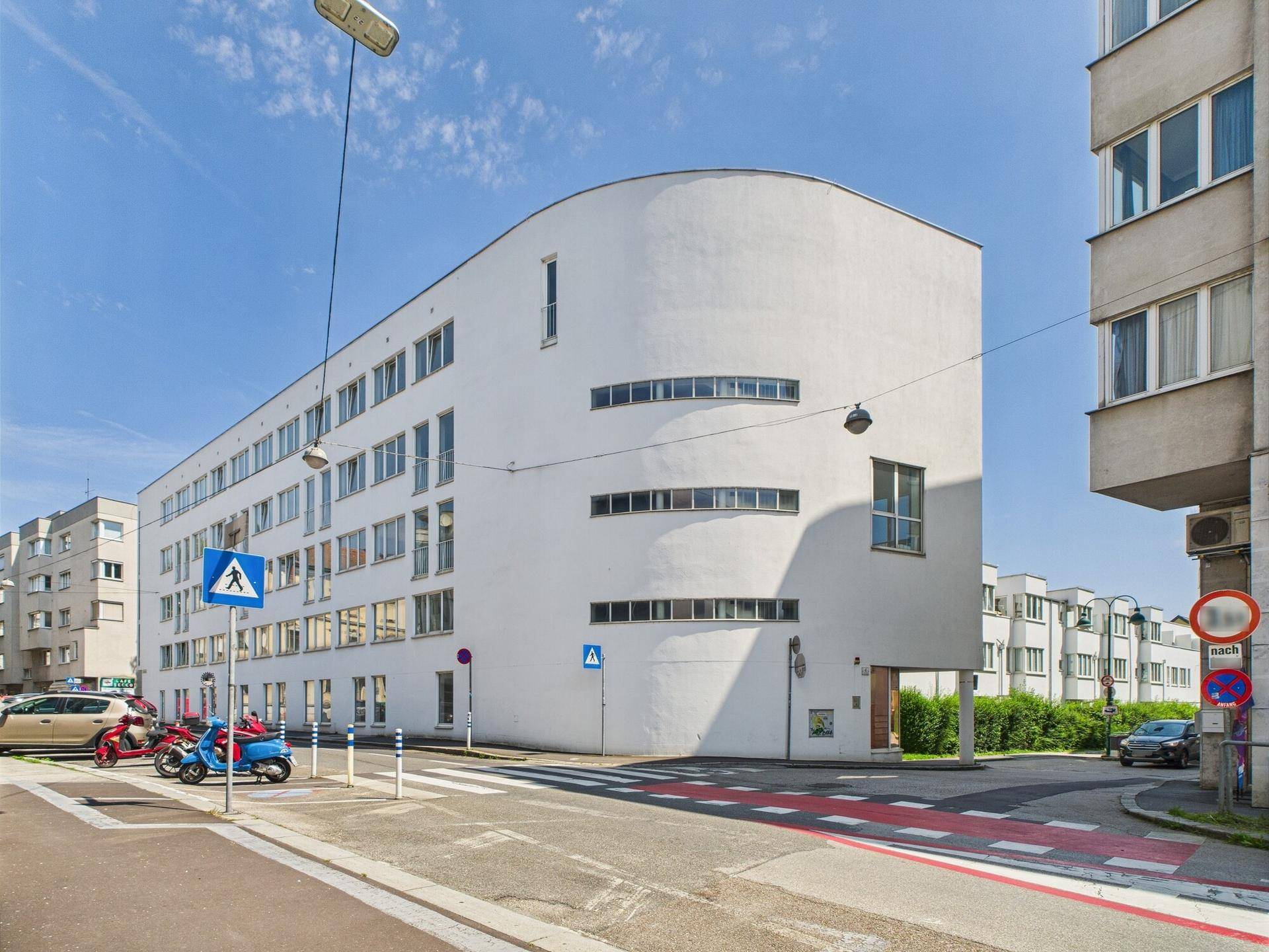Moderne, weiße Gebäudefassade mit geschwungener Architektur an einer Straßenecke unter blauem Himmel.