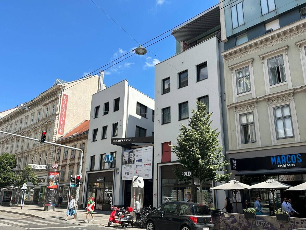 Straßenansicht eines Gebäudekomplexes mit Geschäften im Erdgeschoss und belebter Umgebung.