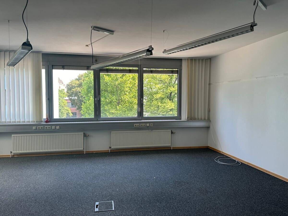 Geräumiges, leeres Büro mit dunklem Teppichboden und großen Fenstern mit Blick ins Grüne.