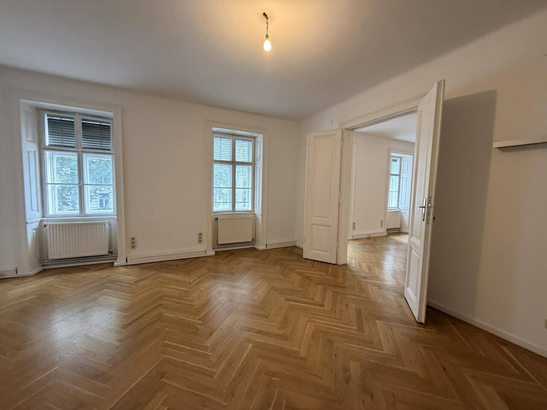 Heller Wohnraum mit Fischgrätparkett, hohen Fenstern und einem Blick in den angrenzenden Raum.
