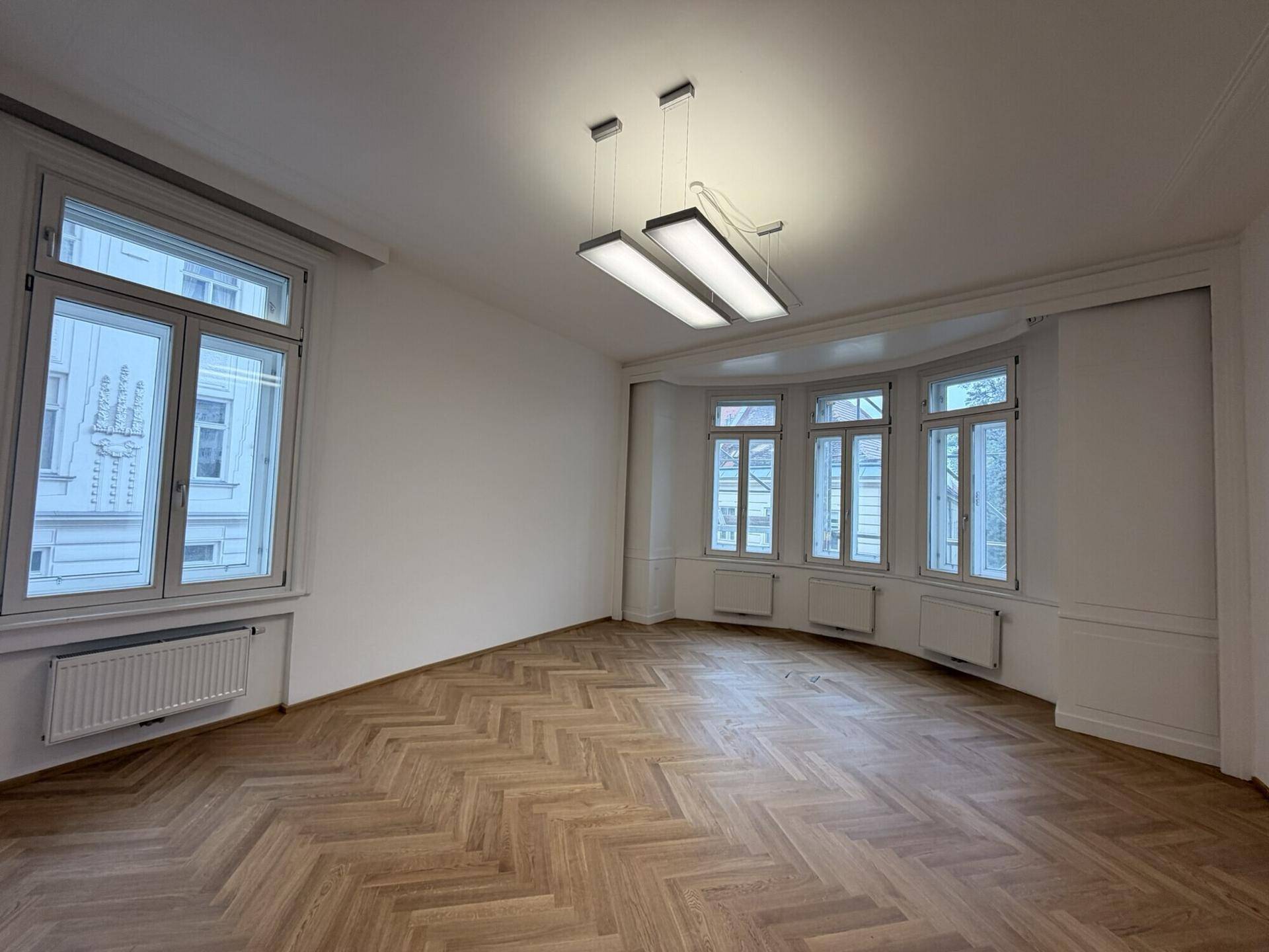 Geräumiger, heller Raum mit Fischgrätparkett, großen Fenstern und einem Erker, der viel Licht bietet.
