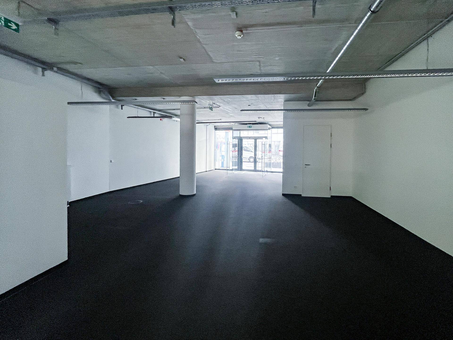 Weitläufiger Gewerberaum mit dunklem Teppichboden, hohen Decken und großen Eingangstüren, geeignet für verschiedene Nutzungen.