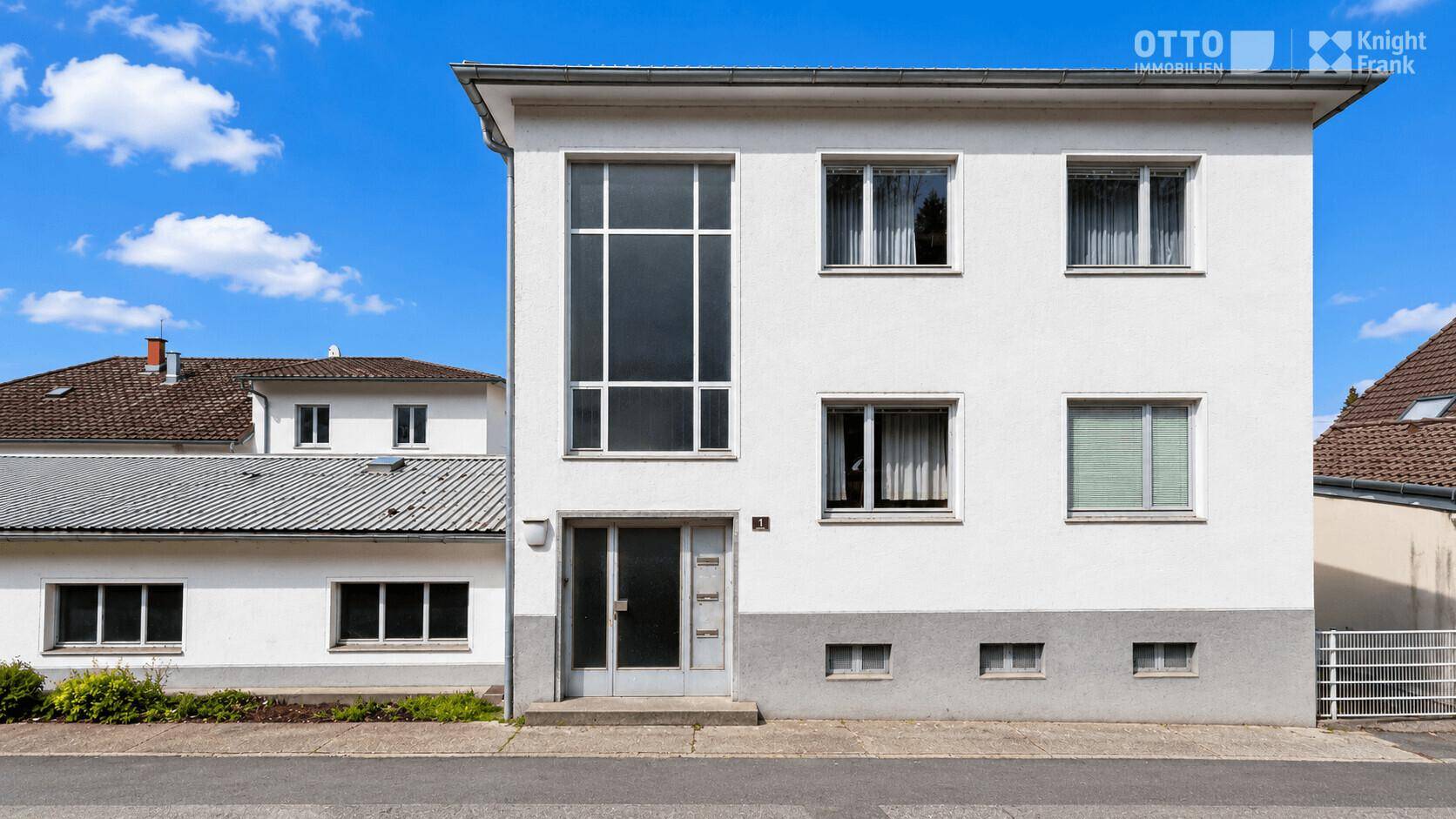 Vorderansicht eines weißen Gebäudes mit einer markanten hohen Fensterfront und weiteren Fenstern.