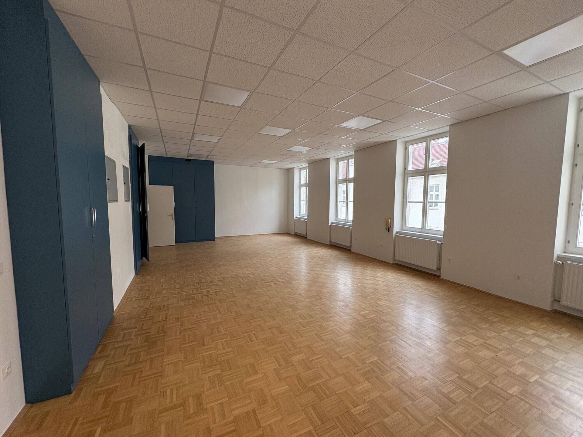 Geräumiger Büroraum mit Parkettboden, blauen Einbauschränken und mehreren Fenstern.