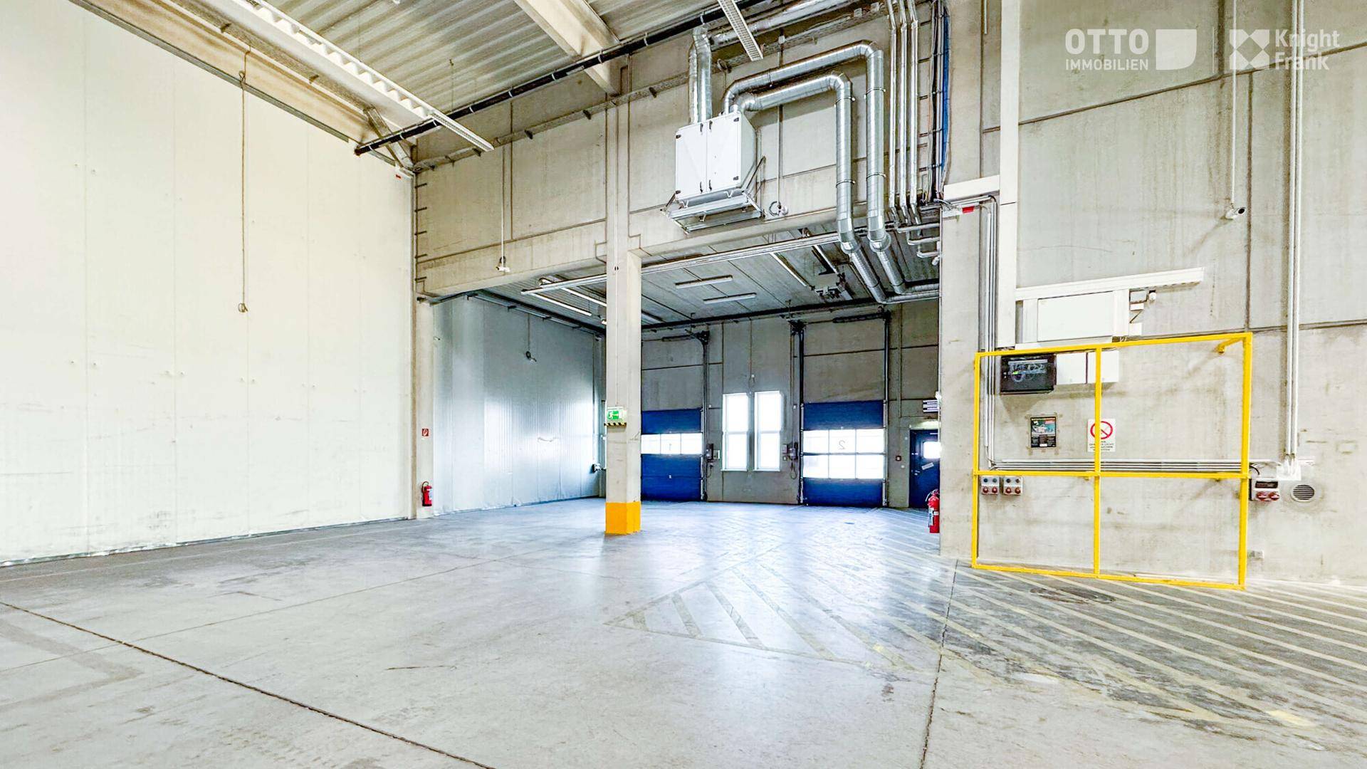 Industrielle Lagerhalle mit hohen Decken, sichtbaren Rohren und einem Betonboden mit Markierungen.