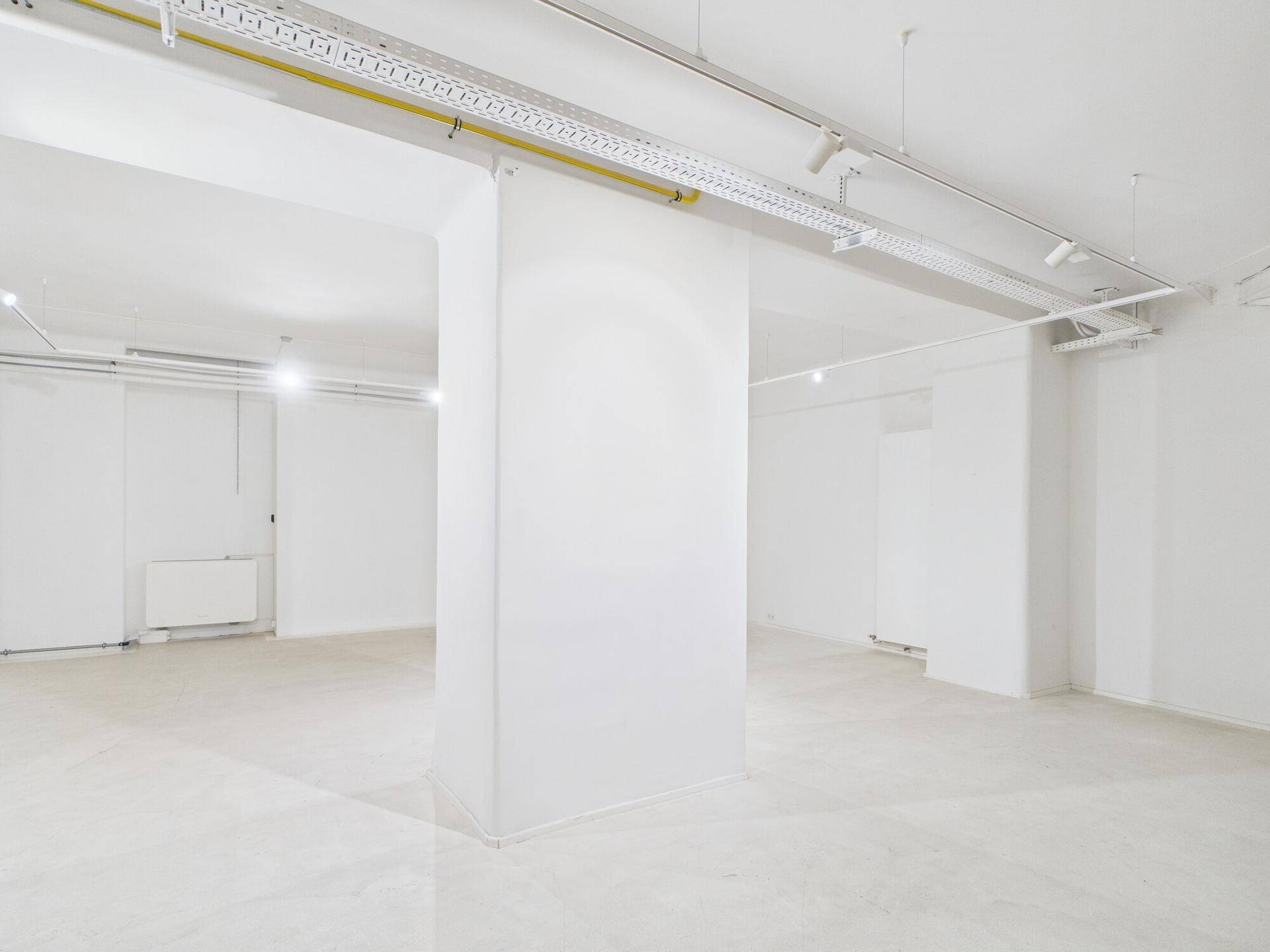 Geräumiger, leerer Gewerberaum mit weißen Wänden, hellem Fliesenboden und sichtbaren Deckeninstallationen.