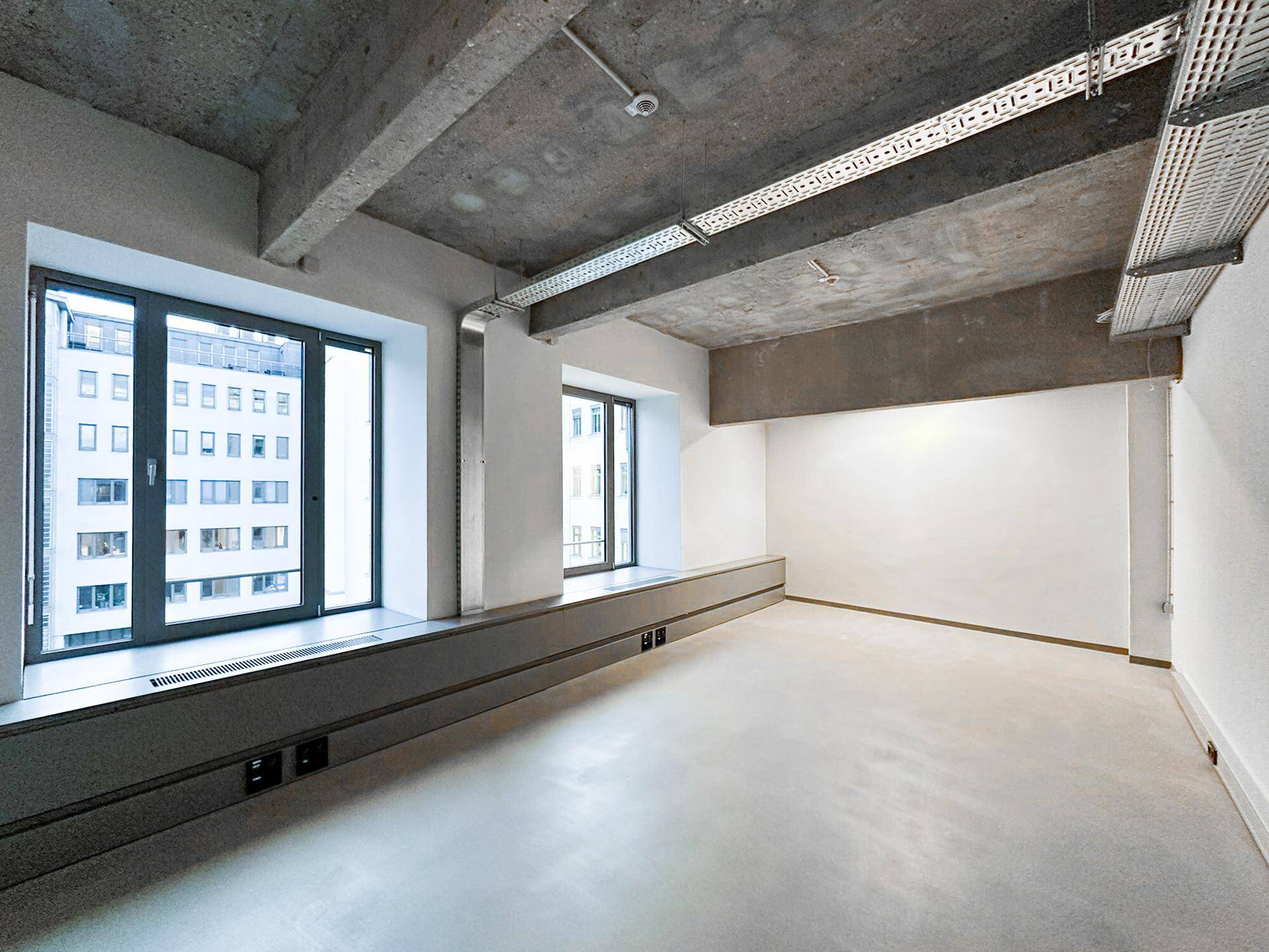 Heller Büroraum mit großen Fenstern, die einen Blick nach draußen ermöglichen, und einer Sichtbetondecke im Loft-Stil.