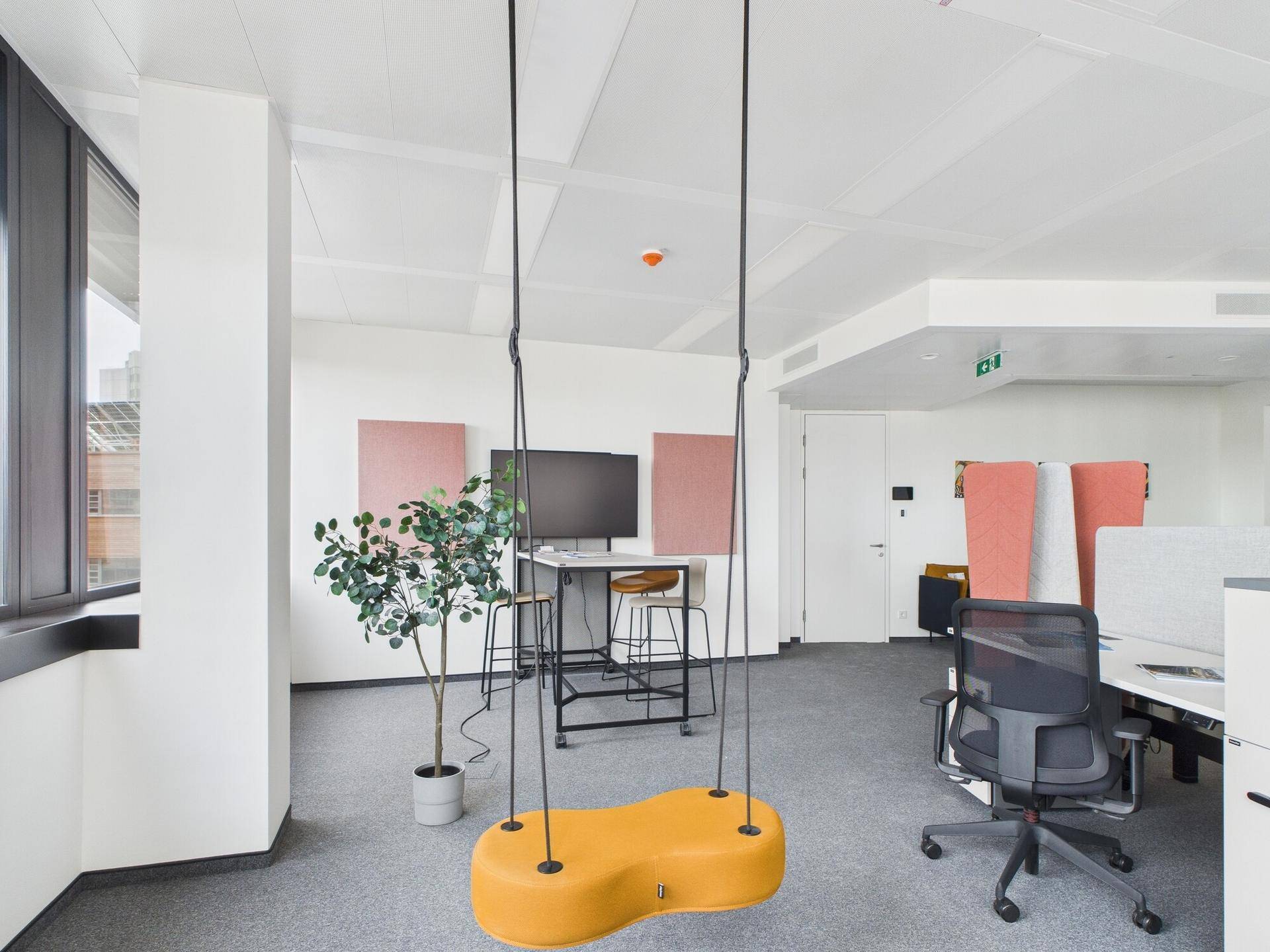 Offenes Büro mit kreativem Schaukelstuhl, modernen Arbeitsplätzen und großen Fenstern, die viel Tageslicht hereinlassen.