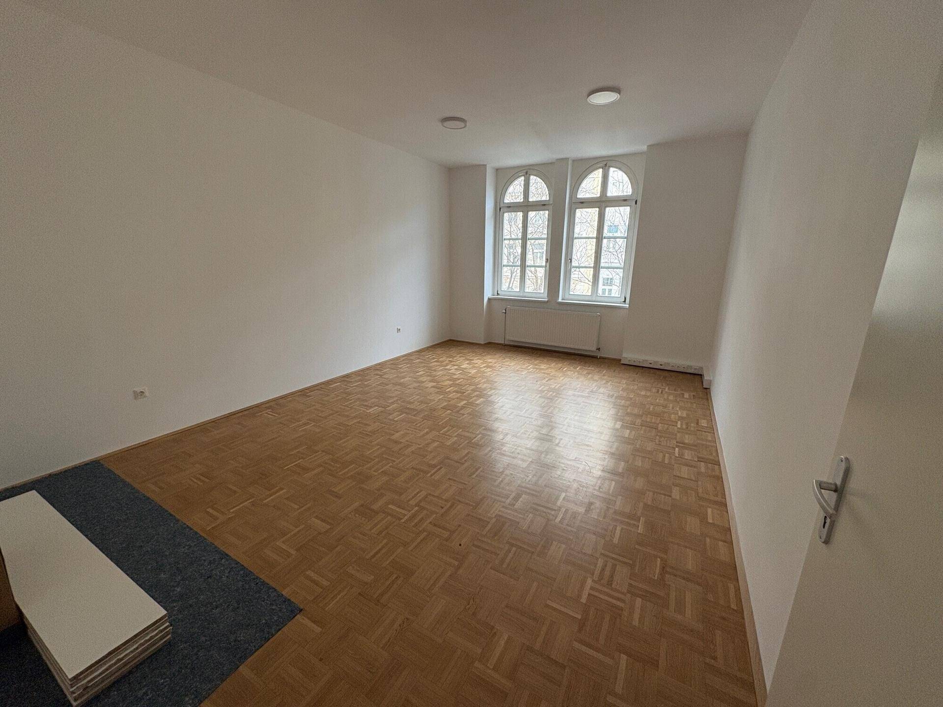 Geräumiger Raum mit Parkettboden und zwei Fenstern, teilweise mit Teppich und Baumaterialien.