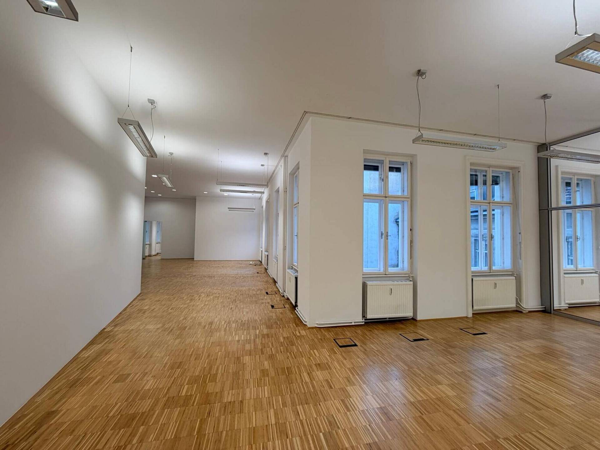 Weitläufiger Büroraum mit Parkettboden, vielen Fenstern und Spiegelwand, der eine helle und offene Arbeitsatmosphäre schafft.
