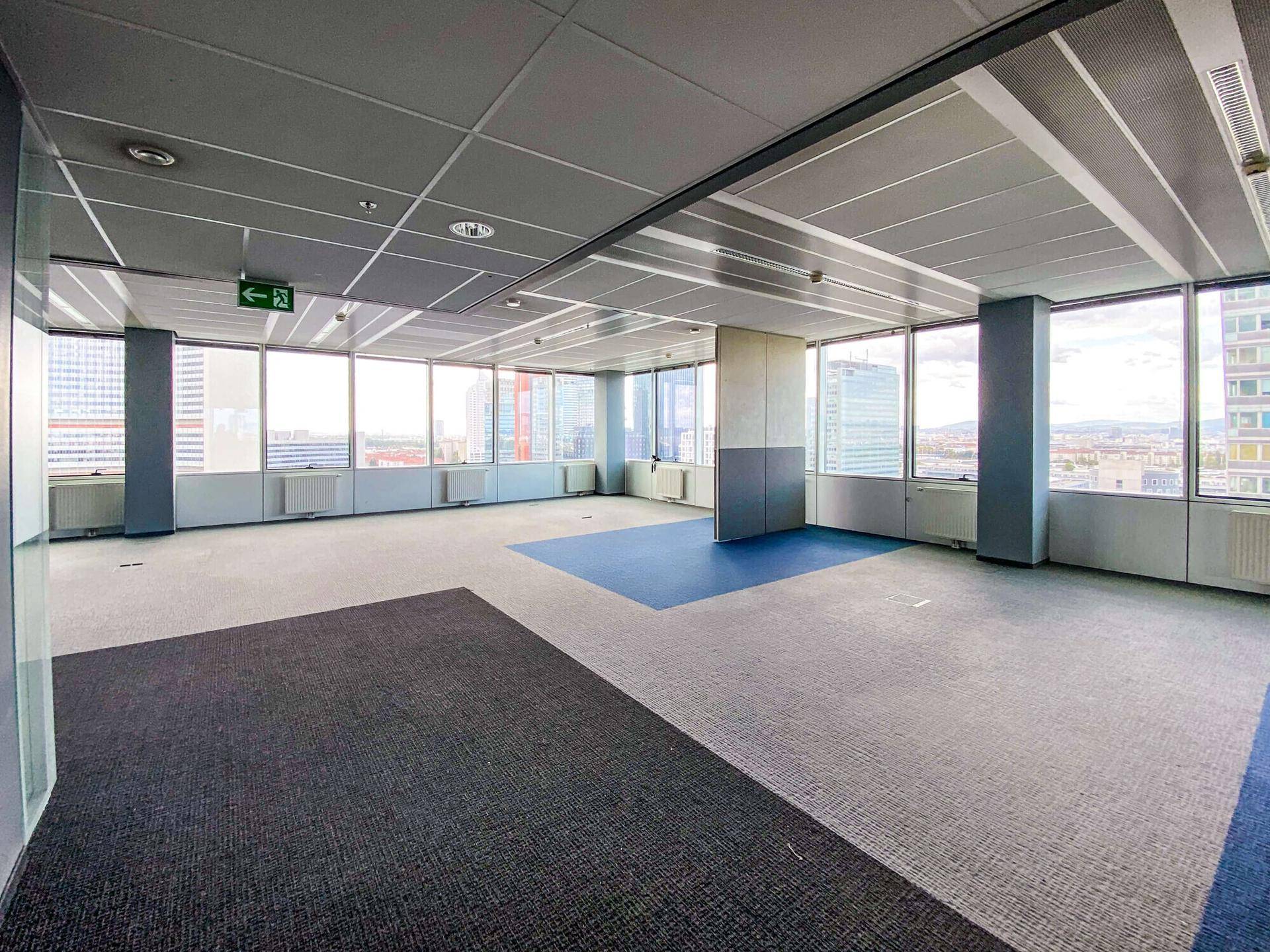 Weitläufiger Bürobereich mit Teppichboden in verschiedenen Farben und großen Fenstern mit Stadtblick.