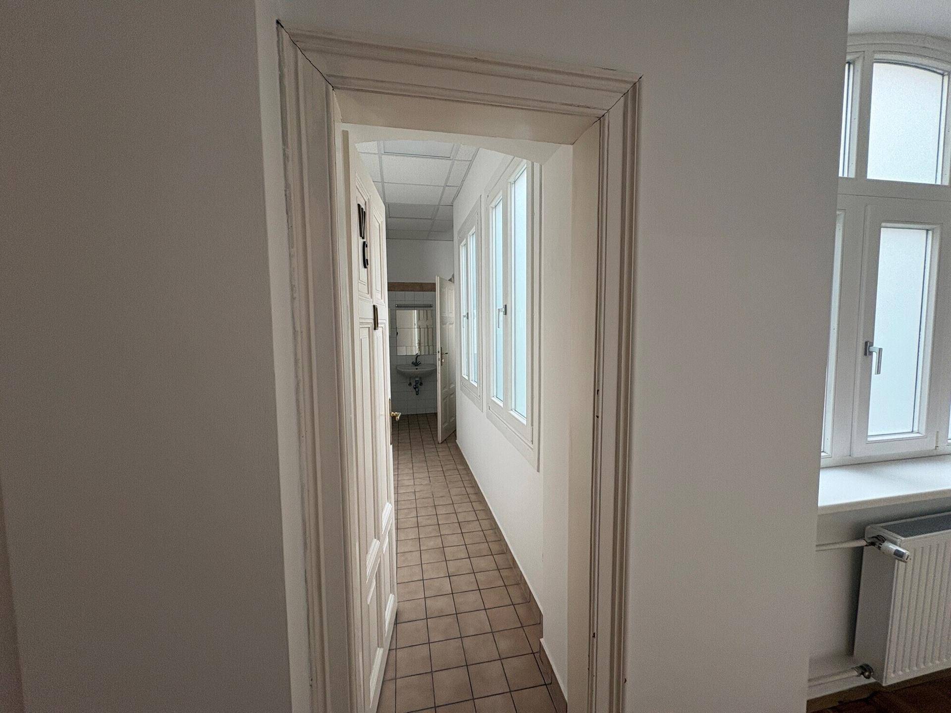 Blick durch eine Tür in einen gefliesten Flur mit Waschbecken und Fenster, mit weißen Wänden.
