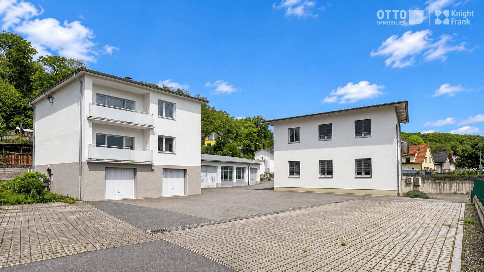 Ansicht von zwei weißen Gebäuden mit Balkonen, Garagen und großzügigen gepflasterten Flächen vor grünen Hügeln.