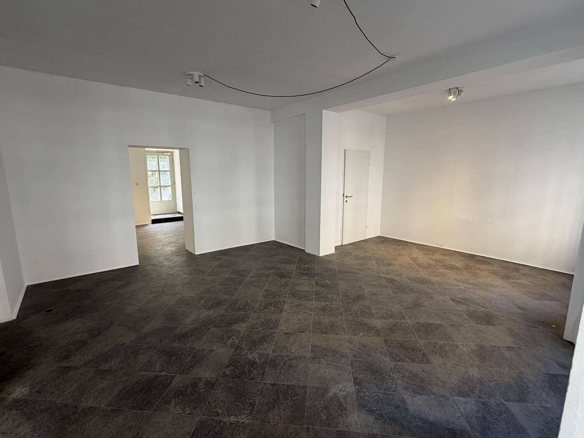 Geräumiger, leerer Innenraum mit dunklem Fliesenboden und einem Durchgang zu einem weiteren Bereich.