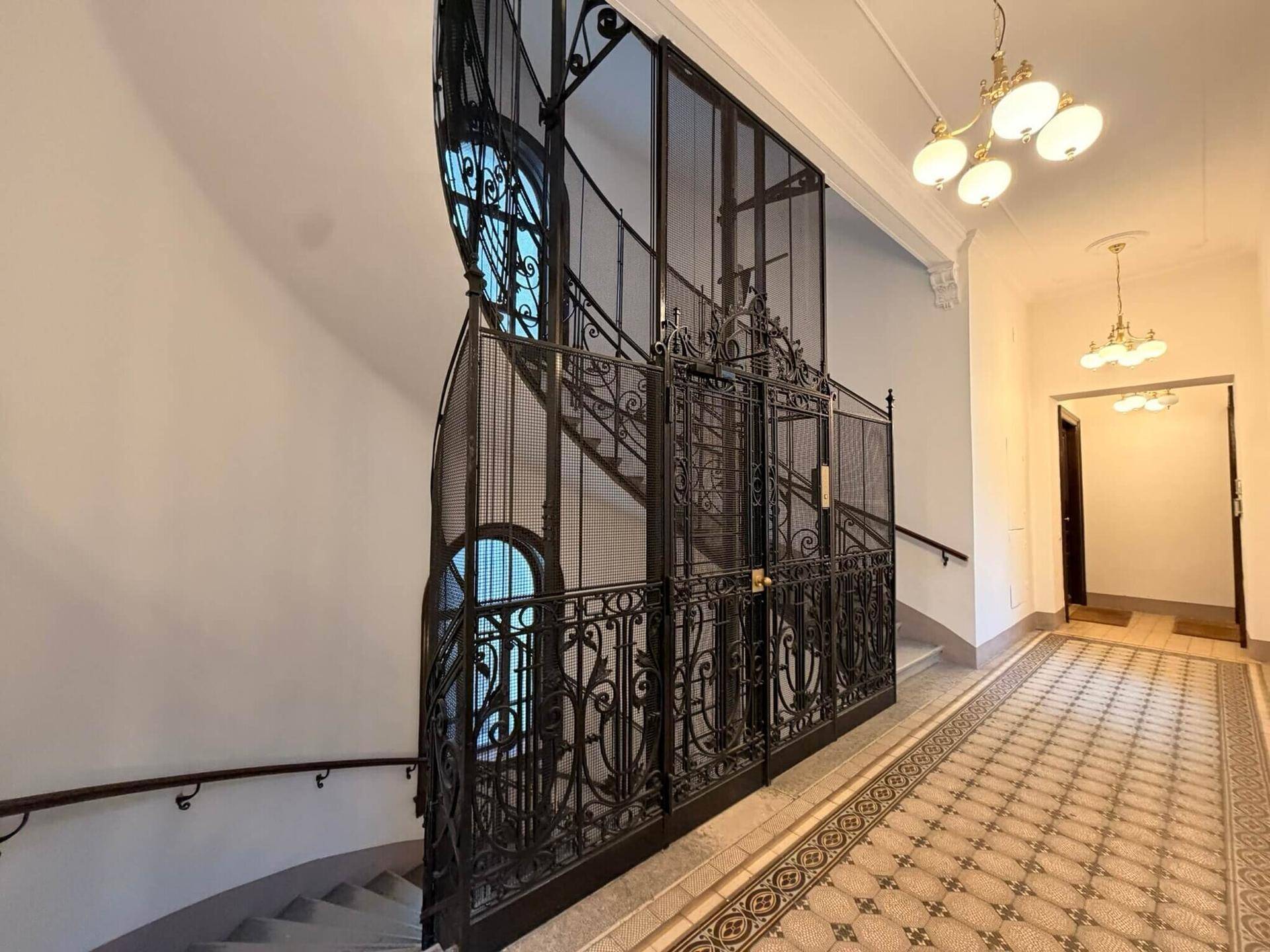 Historisches Treppenhaus mit kunstvollem schmiedeeisernem Aufzug und gemustertem Fliesenboden, das den Charme des Altbaus bewahrt.