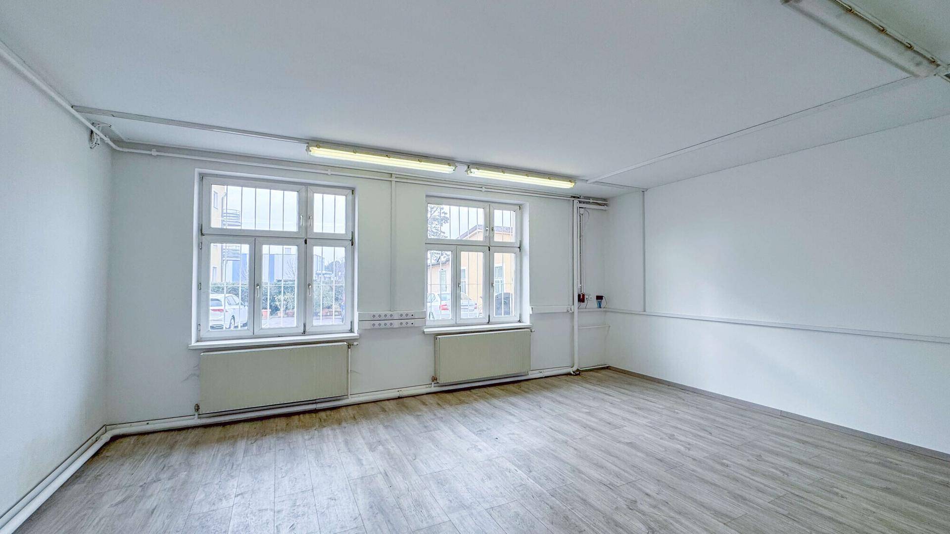 Heller Büroraum mit zwei großen Fenstern, Holzboden und weißen Wänden, ausgestattet mit Heizkörpern.