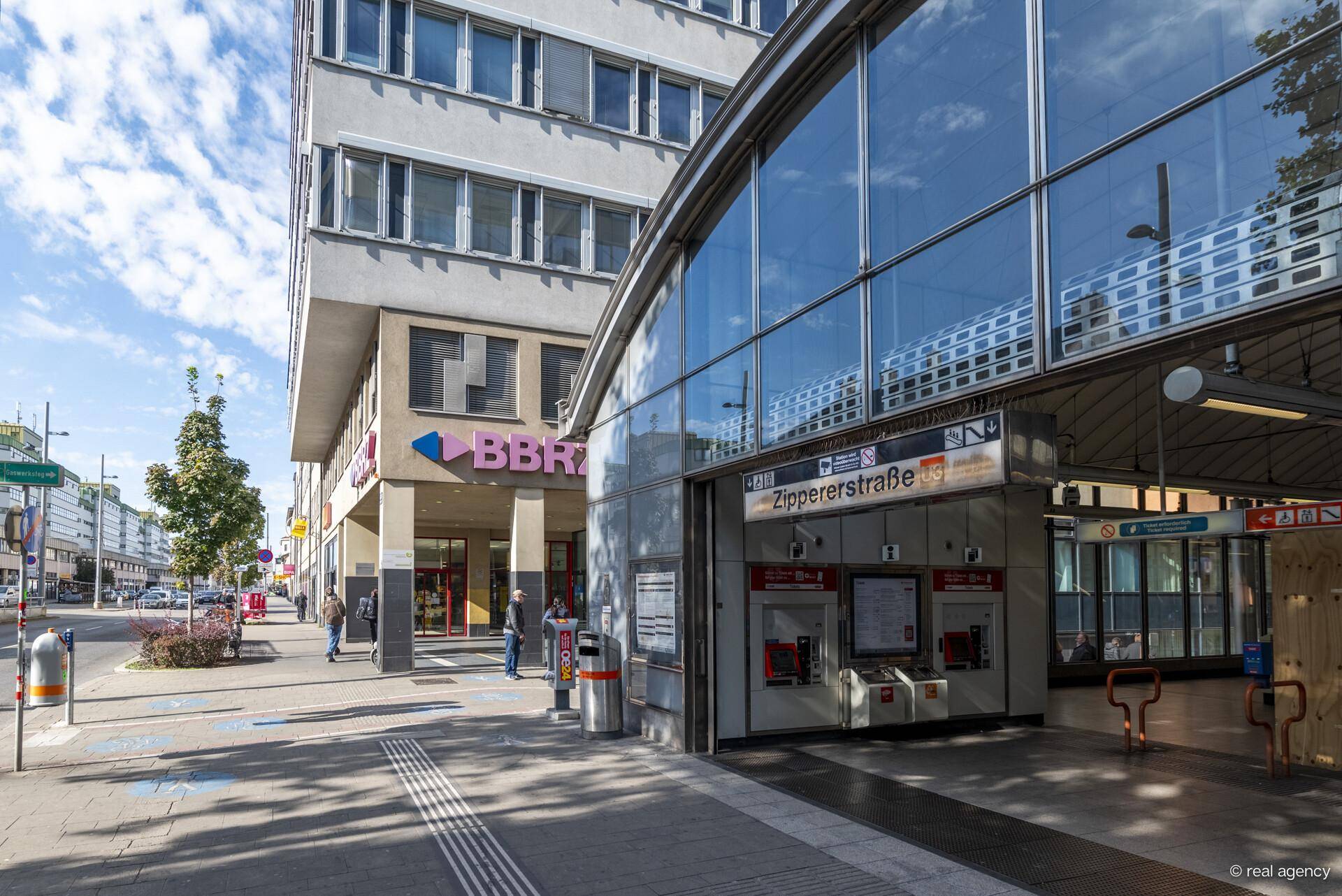 Eingangsbereich eines Gebäudes neben einer U-Bahn-Station mit Glasfassade und Passanten.