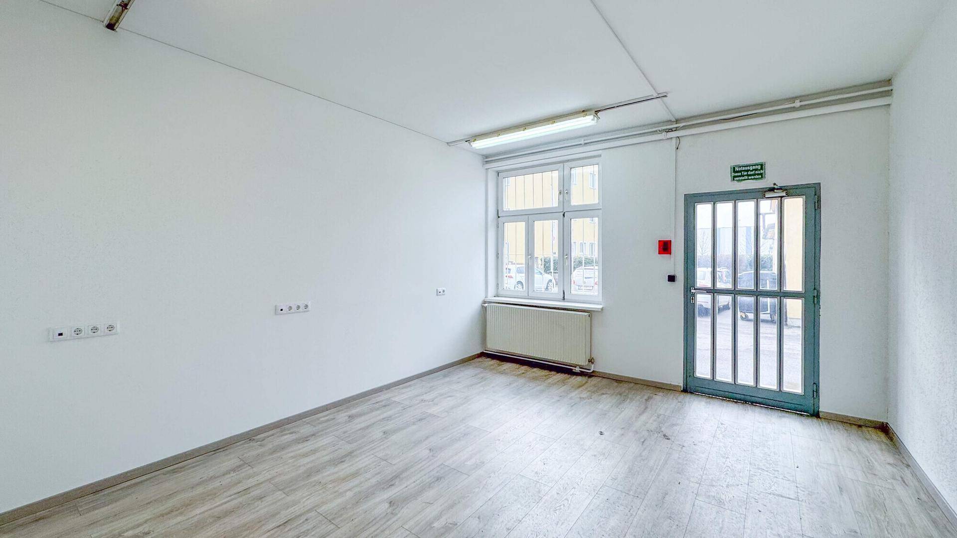 Kleiner, heller Raum mit Holzboden, einem Fenster und einer Außentür mit Gitter, ausgestattet mit Heizkörper.