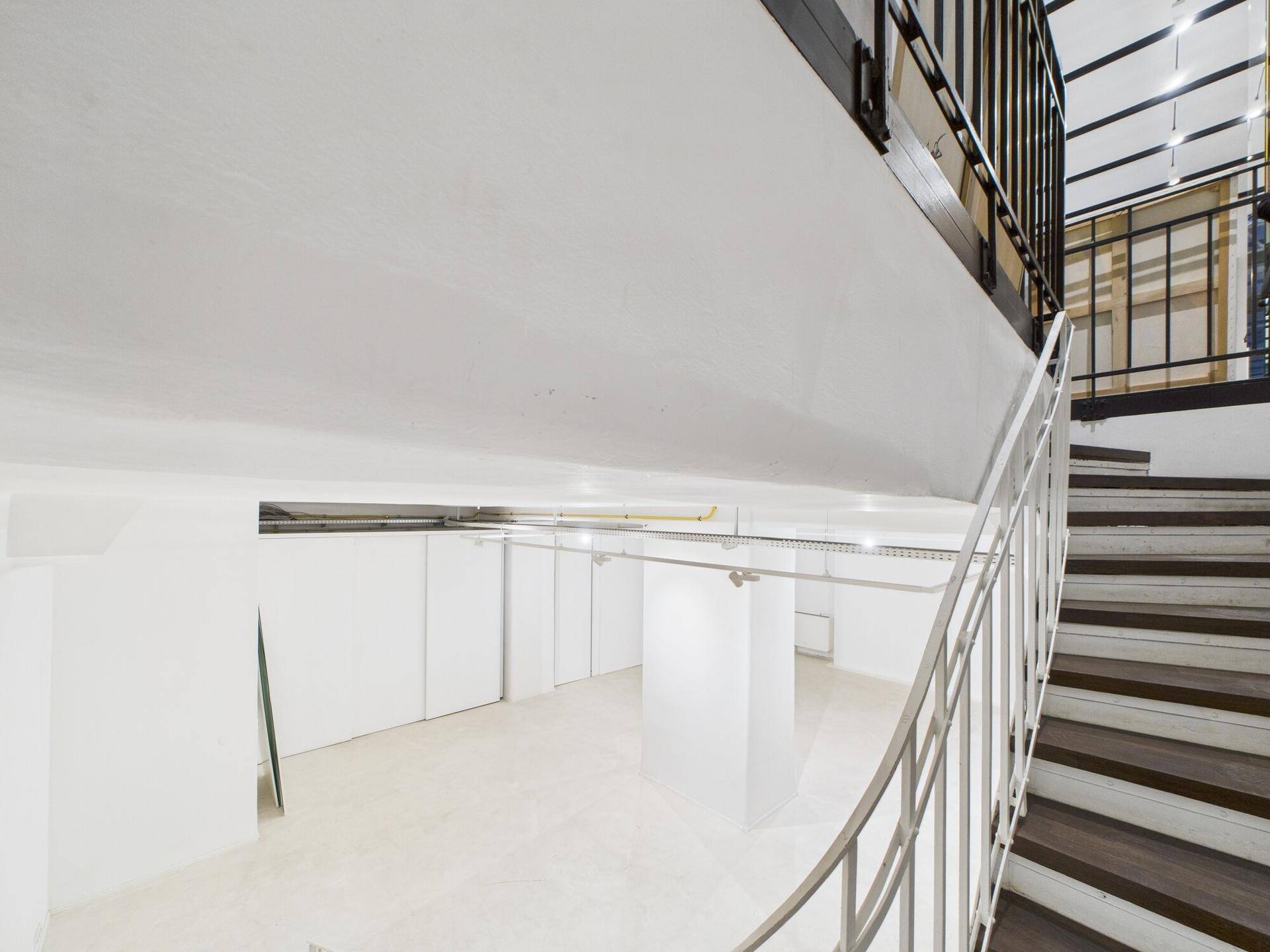 Blick von der Treppe in einen hellen Untergeschoss-Gewerberaum mit weißen Oberflächen und sichtbaren Leitungen.