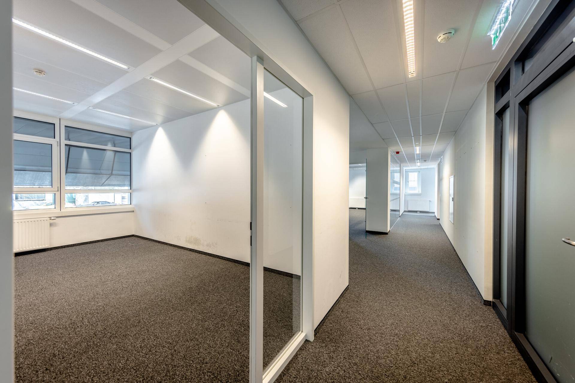 Heller Büroflur mit Teppichboden und Glaswänden, der zu einem leeren Büroraum führt.