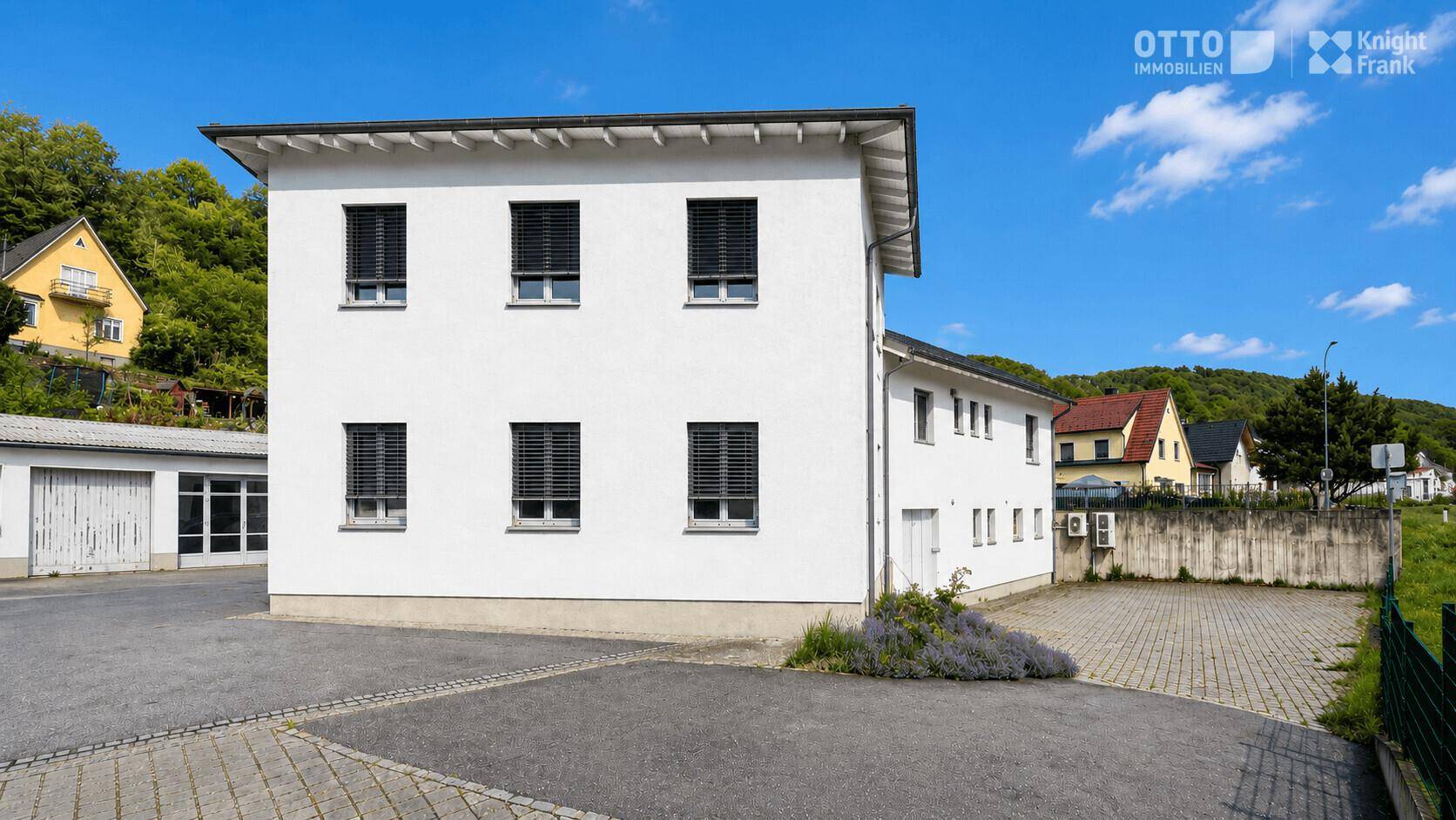 Seitenansicht eines weißen Gebäudes mit schwarzen Fensterläden, gepflastertem Hof und einer kleinen Lavendelpflanzung.