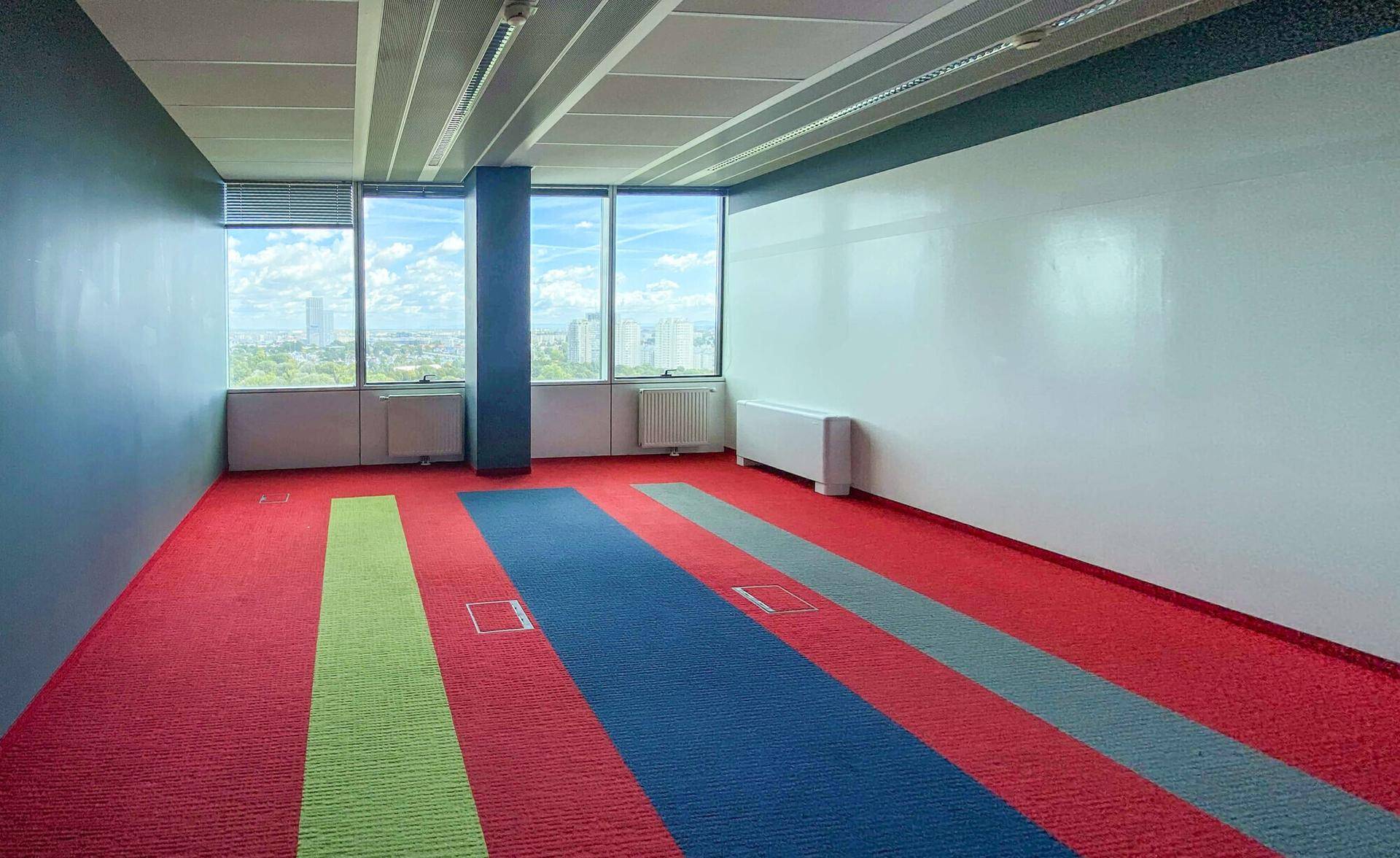 Leeres Büro mit farbigem Teppichboden, weißen Wänden und Fenstern mit Stadtblick.