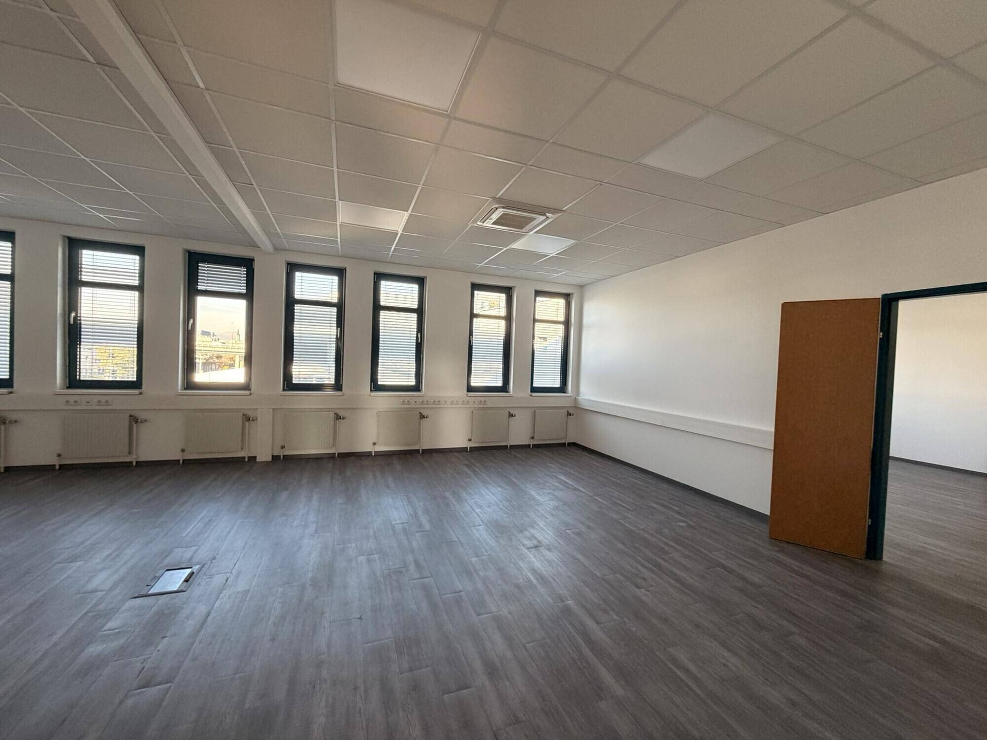 Geräumiger Büroraum mit dunklem Holzboden, weißen Wänden und einer Reihe von Fenstern.