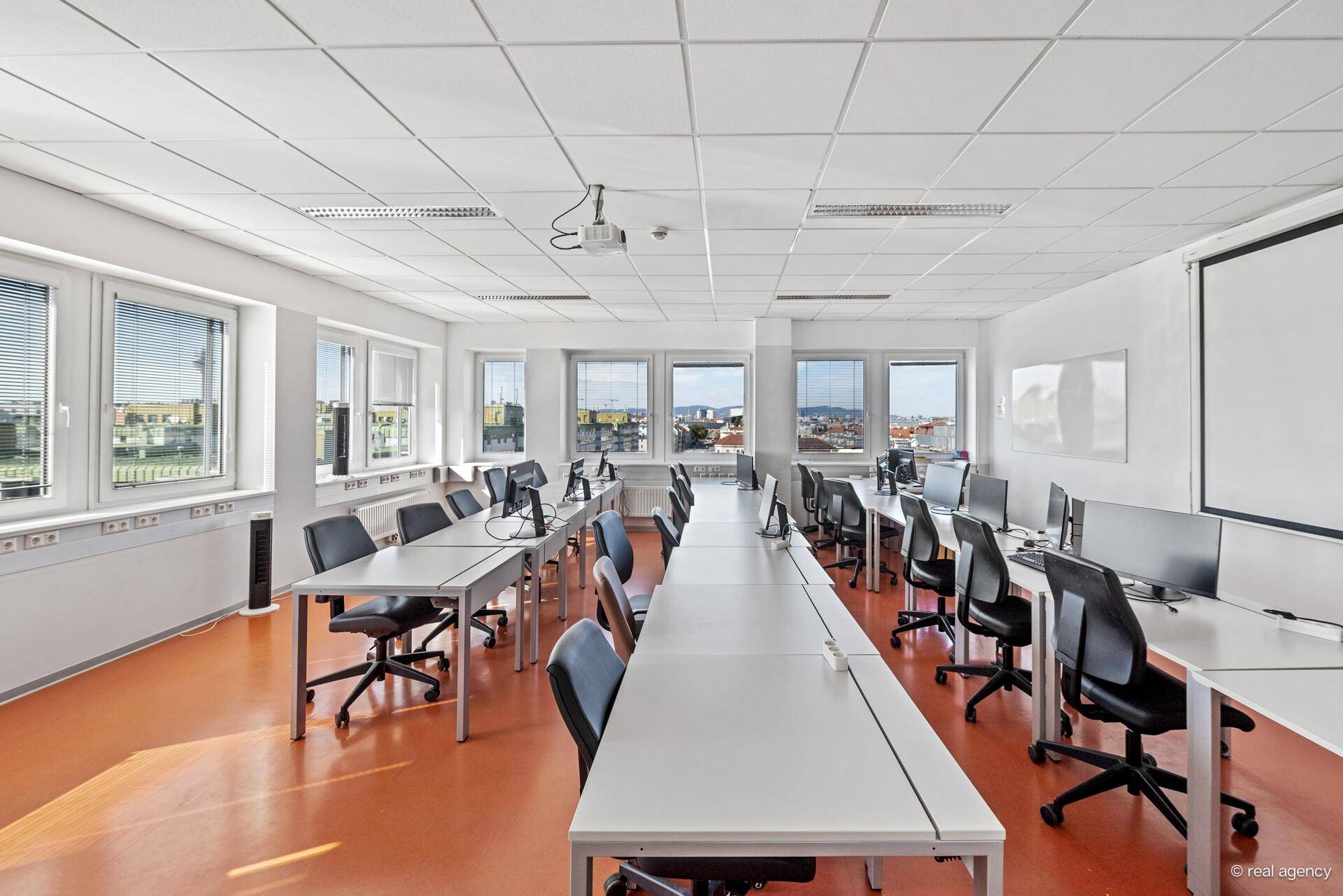 Schulungsraum mit Computern, Tischen, Stühlen und großen Fenstern mit Stadtblick.