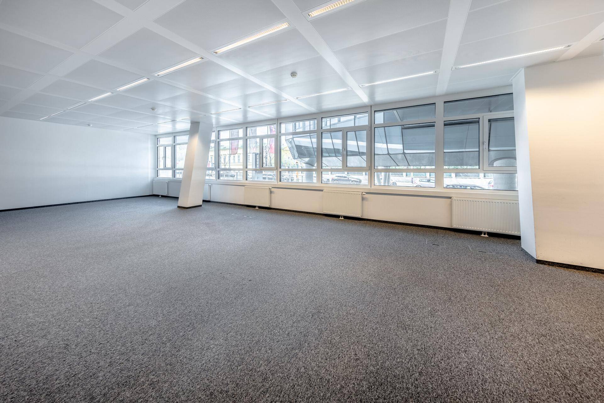 Geräumiger, leerer Büroraum mit Teppichboden, großen Fenstern und Deckenbeleuchtung.