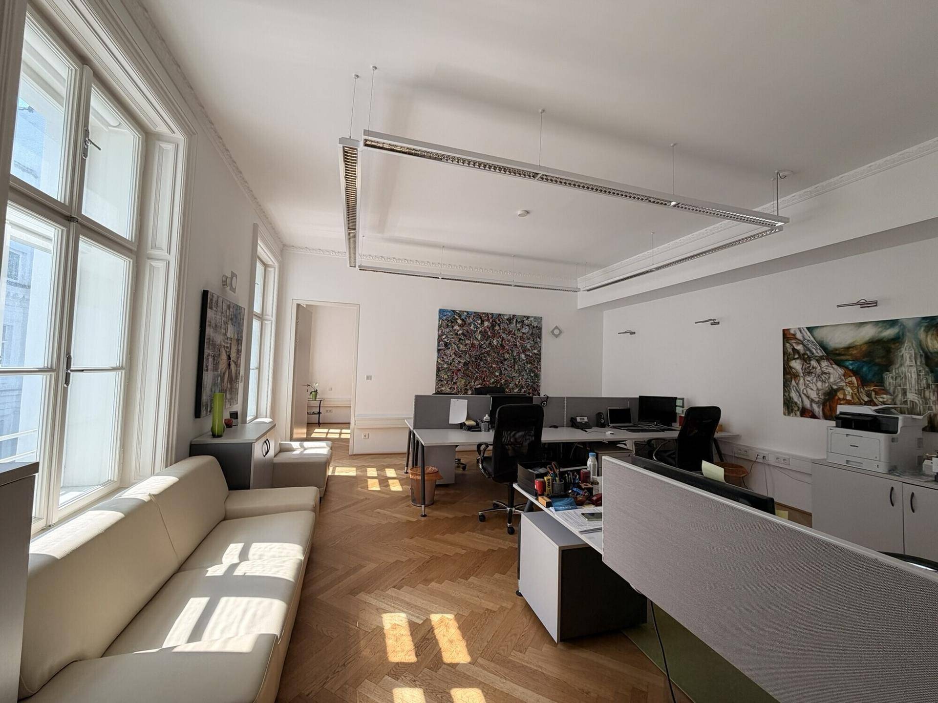 Großes, helles Büro mit mehreren Arbeitsplätzen, Fischgrätparkett und großen Fenstern.