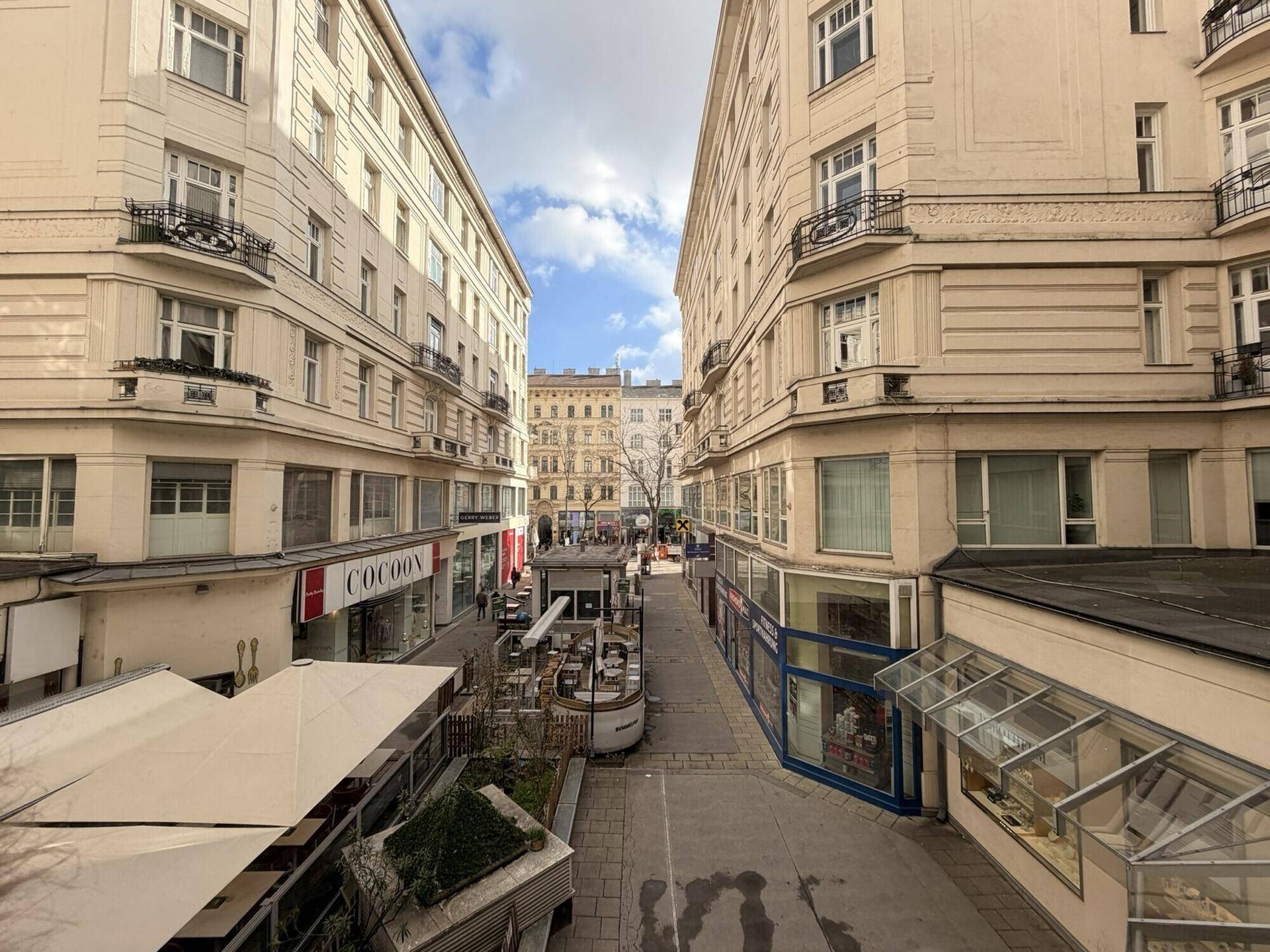 Blick auf eine belebte Gasse zwischen Gebäuden mit Geschäften und Außengastronomiebereichen.