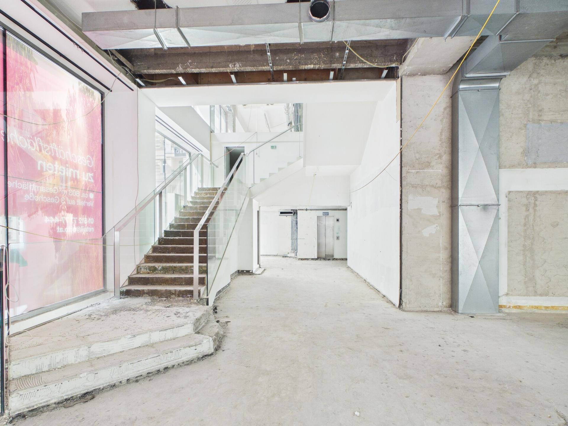 Großzügiger Eingangsbereich eines Gewerbeobjekts im Rohbau mit moderner Treppe und sichtbaren Lüftungskanälen.