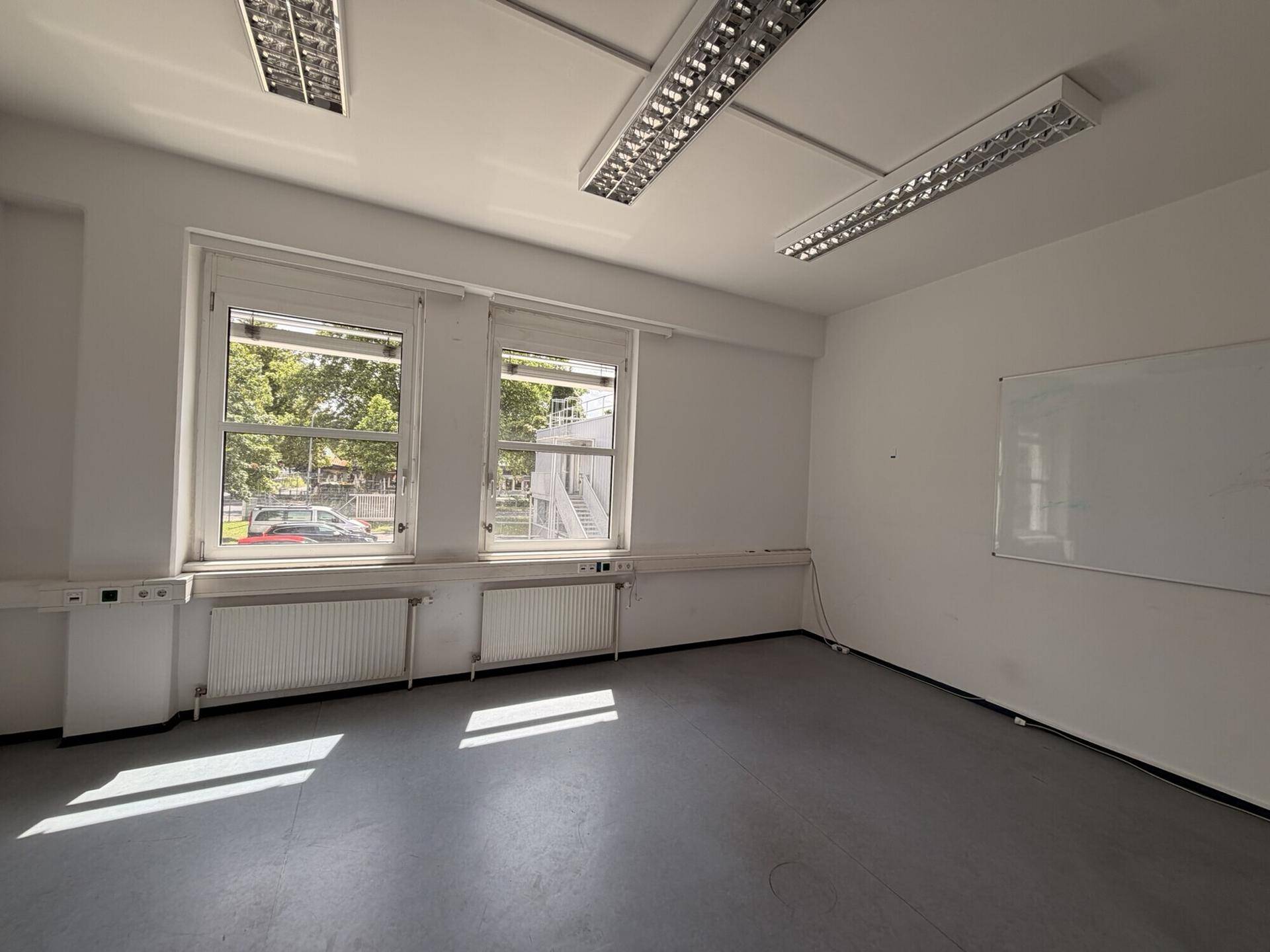Geräumiger Büroraum mit großen Fenstern, Jalousien und einem funktionalen grauen Bodenbelag, ideal für Arbeitsbereiche.