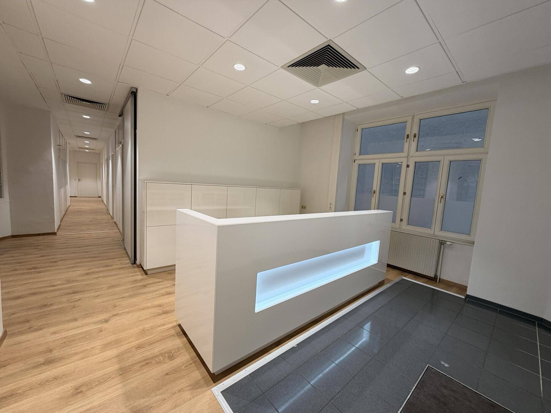 Moderner Empfangsbereich mit beleuchteter Theke, Laminatboden und großen Fenstern.