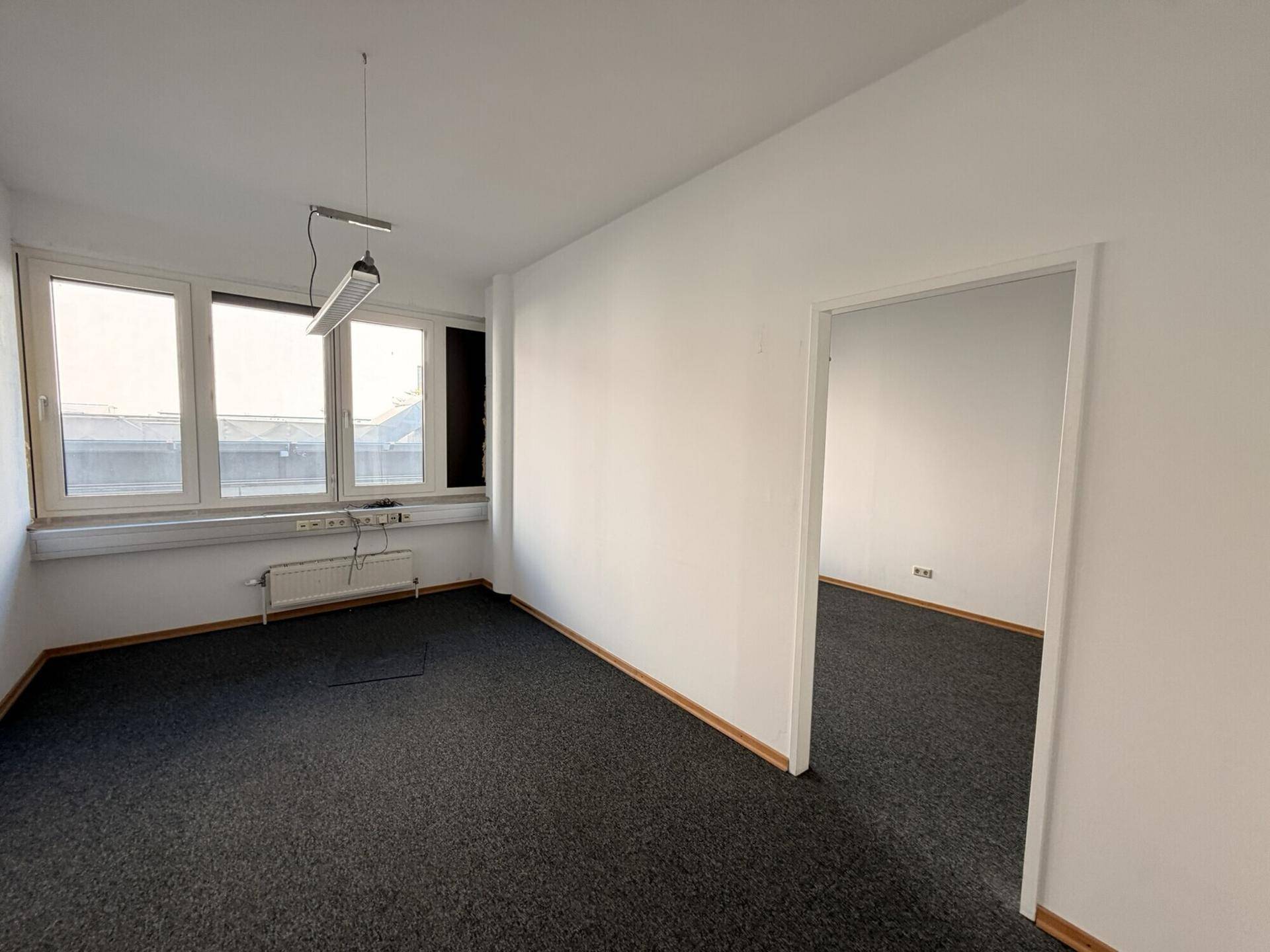 Leeres Büro mit dunklem Teppichboden und Fenstern, das durch eine Tür mit einem weiteren Raum verbunden ist.