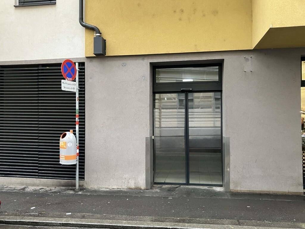 Gebäudeeingang mit großen Glastüren und einer grauen Wand, daneben ein Lüftungsgitter und ein Verkehrsschild.