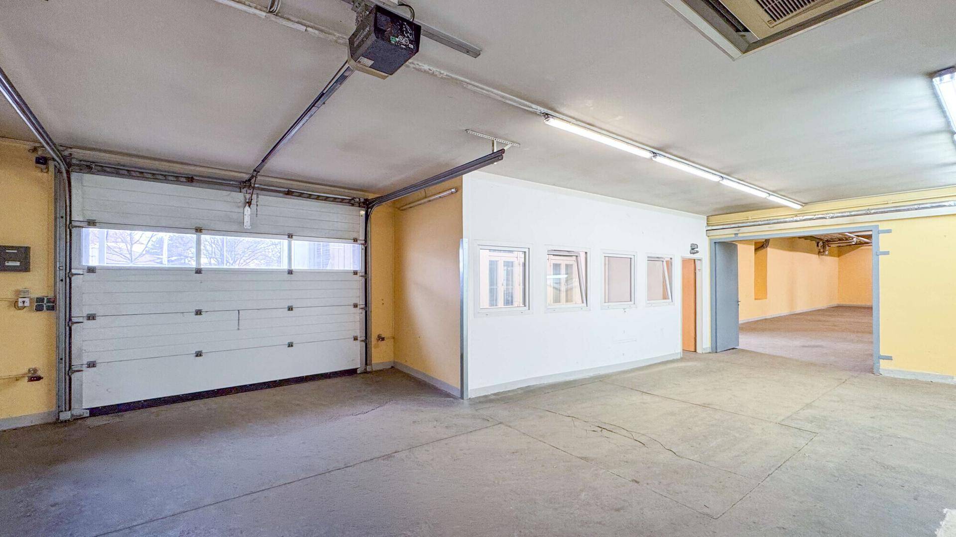 Innenansicht einer Garage mit großem Rolltor, Betonboden und Zugang zu weiteren Räumen, gut beleuchtet.