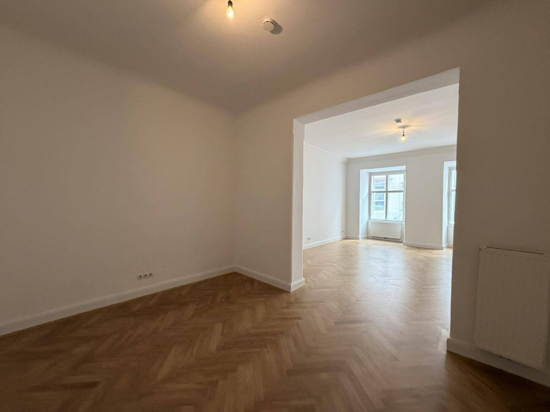 Heller Innenraum mit Fischgrätparkett und einem offenen Durchgang zu einem weiteren Zimmer, das viel Potenzial bietet.