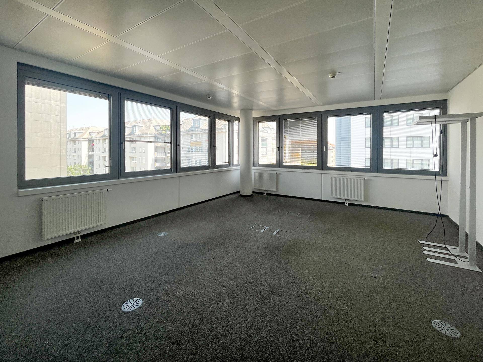 Großes Büro mit vielen Fenstern, Teppichboden und Blick auf umliegende Gebäude, bietet viel Tageslicht.