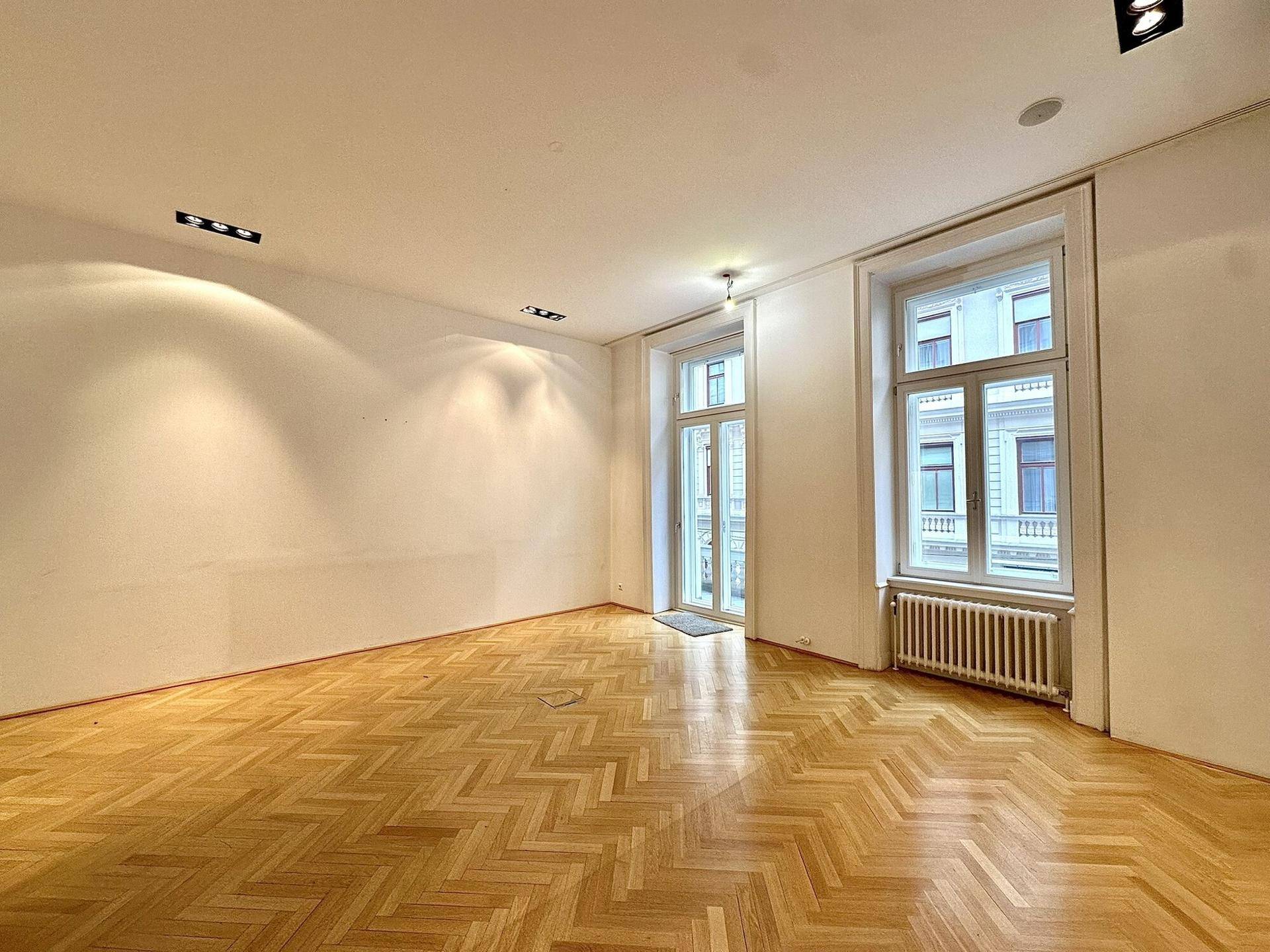 Heller Wohnraum mit Fischgrätparkettboden und hohen Fenstern, die viel Tageslicht hereinlassen.