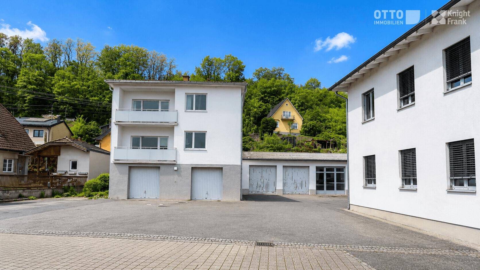 Ansicht von zwei weißen Gebäuden mit Garagen und Balkonen, umgeben von einer grünen Hügellandschaft.