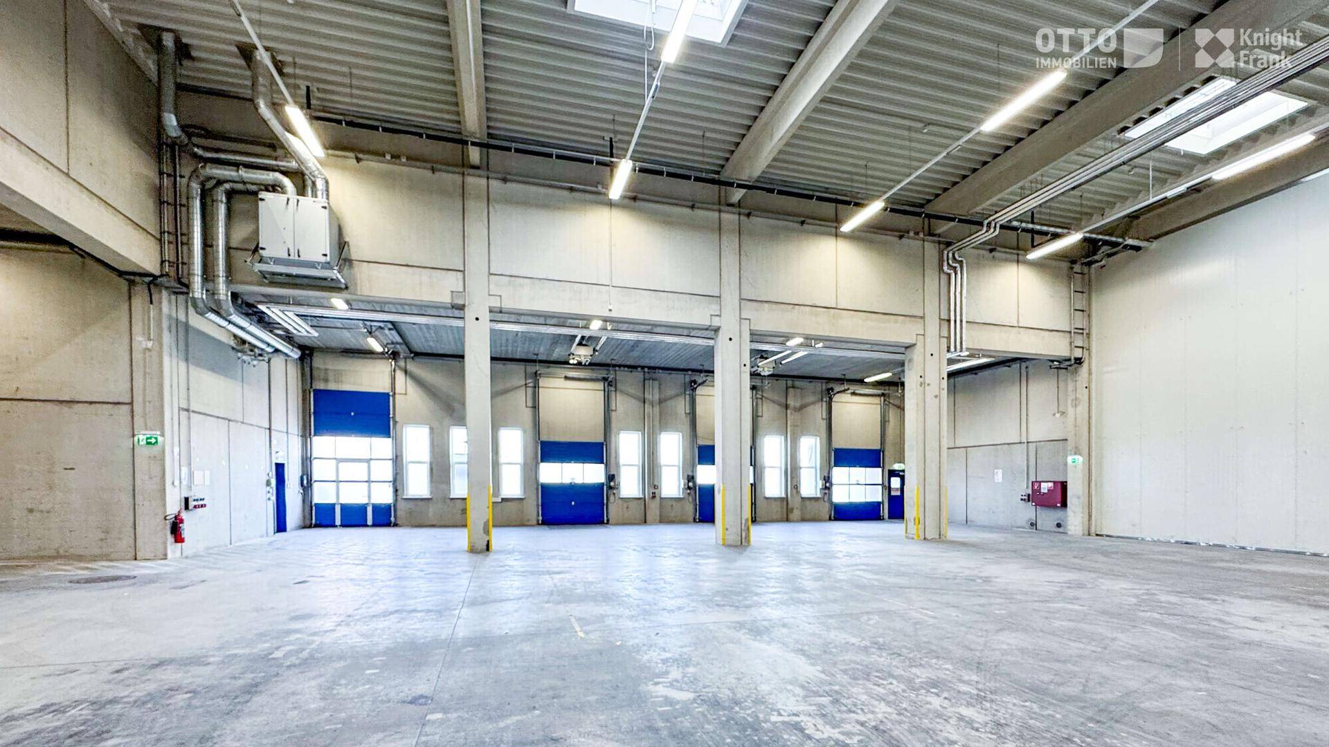 Große Lagerhalle mit mehreren Laderampen, hohen Decken und Betonboden, gut beleuchtet.
