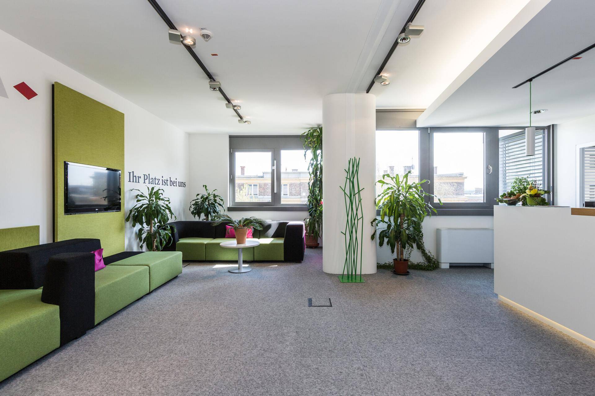 Heller Empfangsbereich mit grünen Sofas, vielen Pflanzen und einem Fernseher an der Wand.