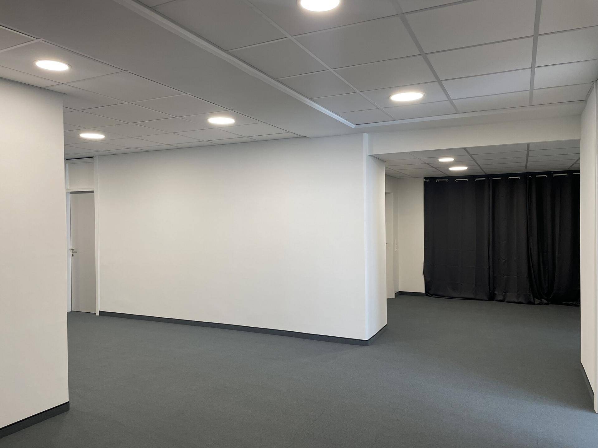 Großer, offener Bürobereich mit Teppichboden, Deckenbeleuchtung und einem abgetrennten Bereich mit Vorhang.