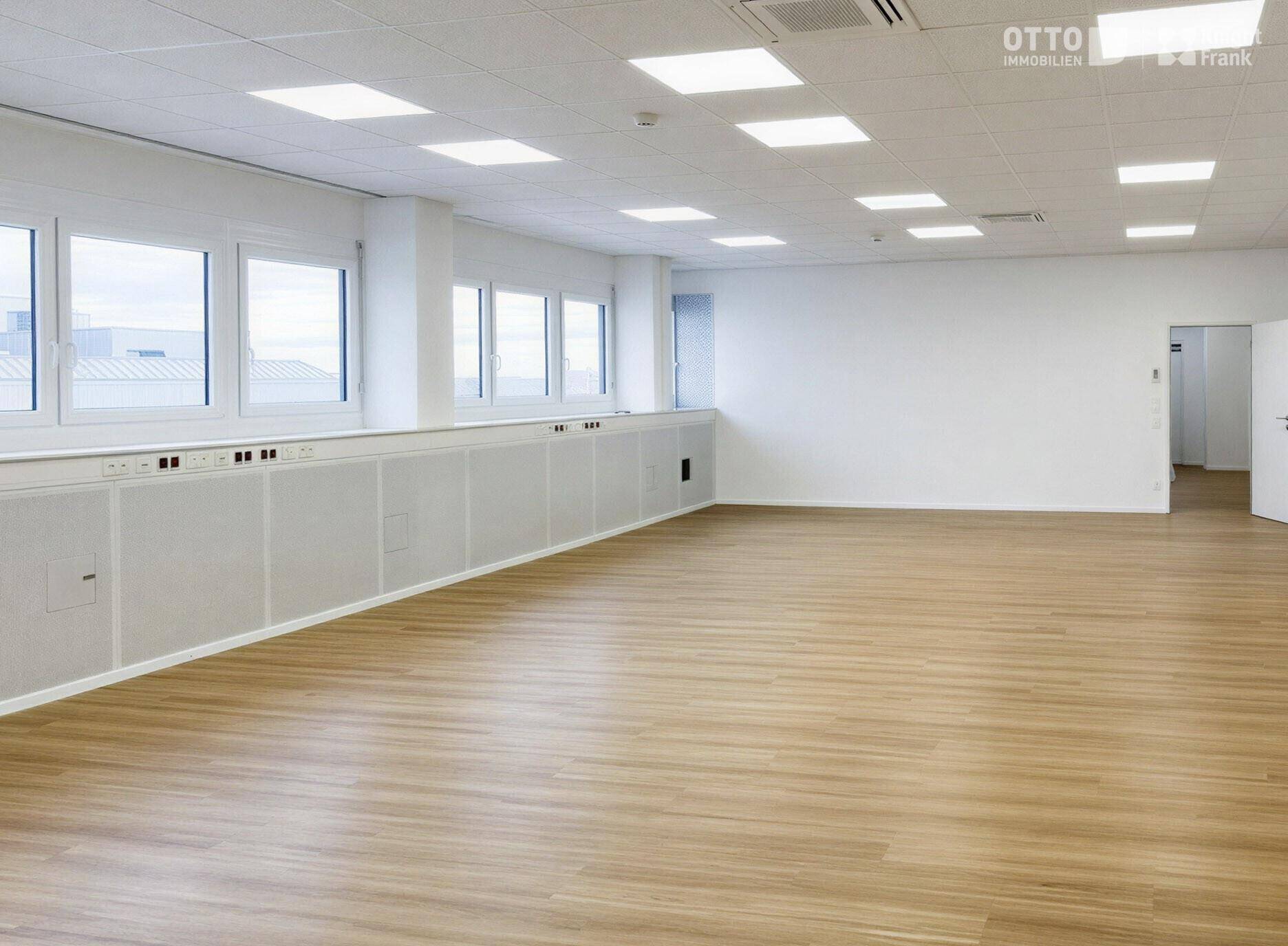 Großes, leeres Büro mit Holzfußboden, vielen Fenstern und moderner Deckenbeleuchtung, bereit zur Einrichtung.