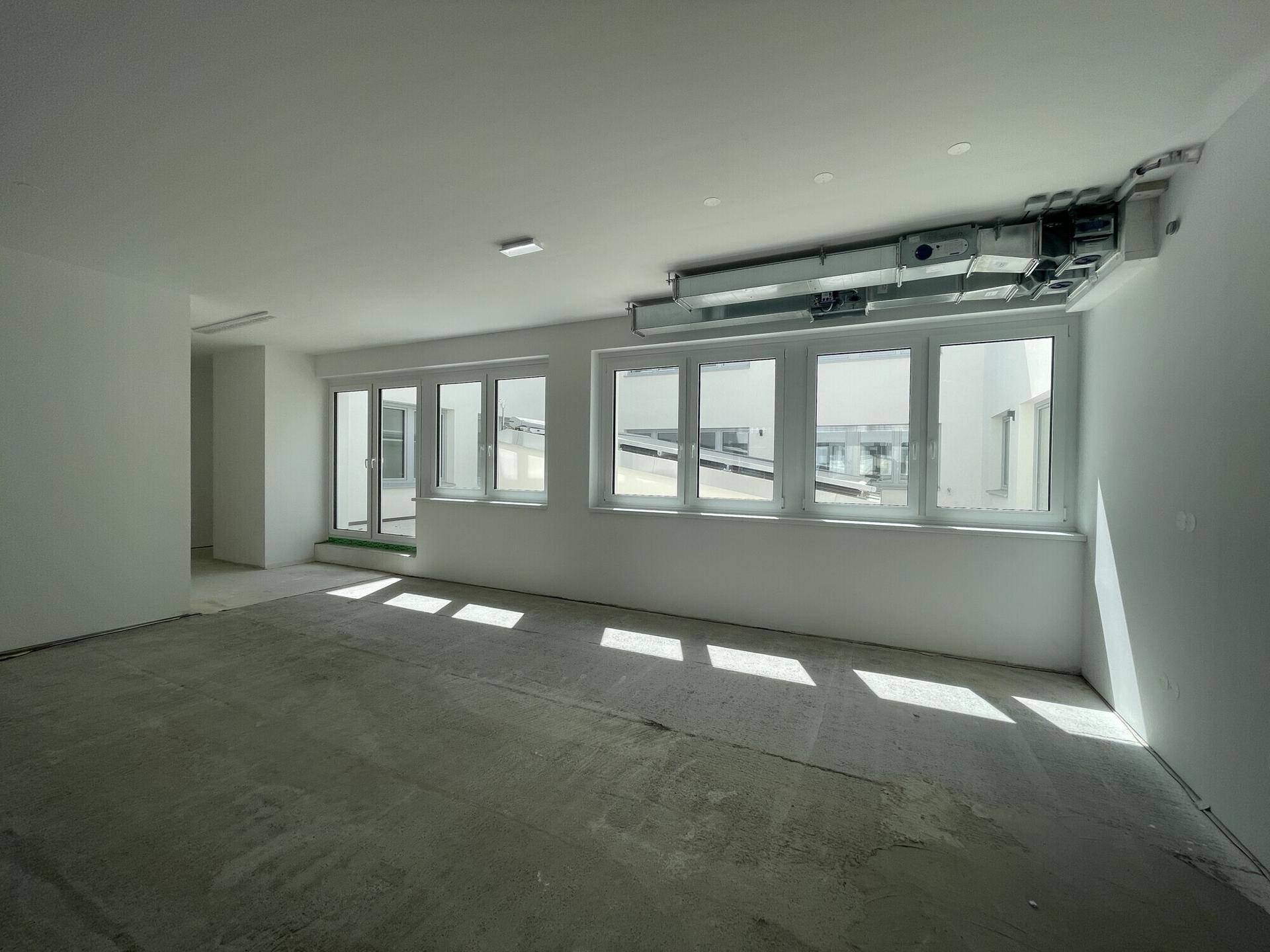 Heller Büroraum im Rohbau mit sichtbaren Lüftungsrohren und Zugang zu einem Außenbereich.