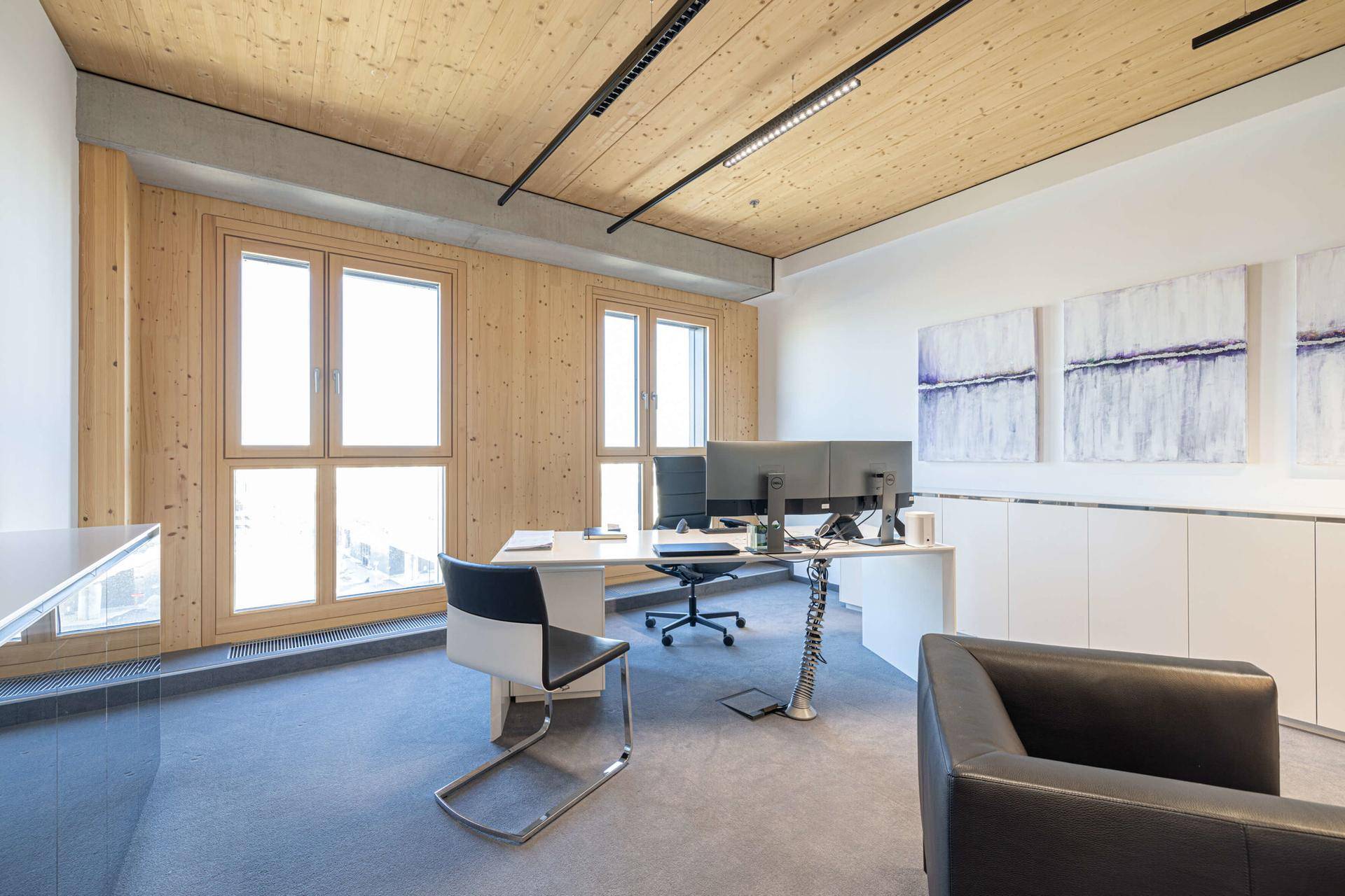Helles und modernes Büro mit Holzwänden, Teppichboden und großen Fenstern, die viel Tageslicht hereinlassen.
