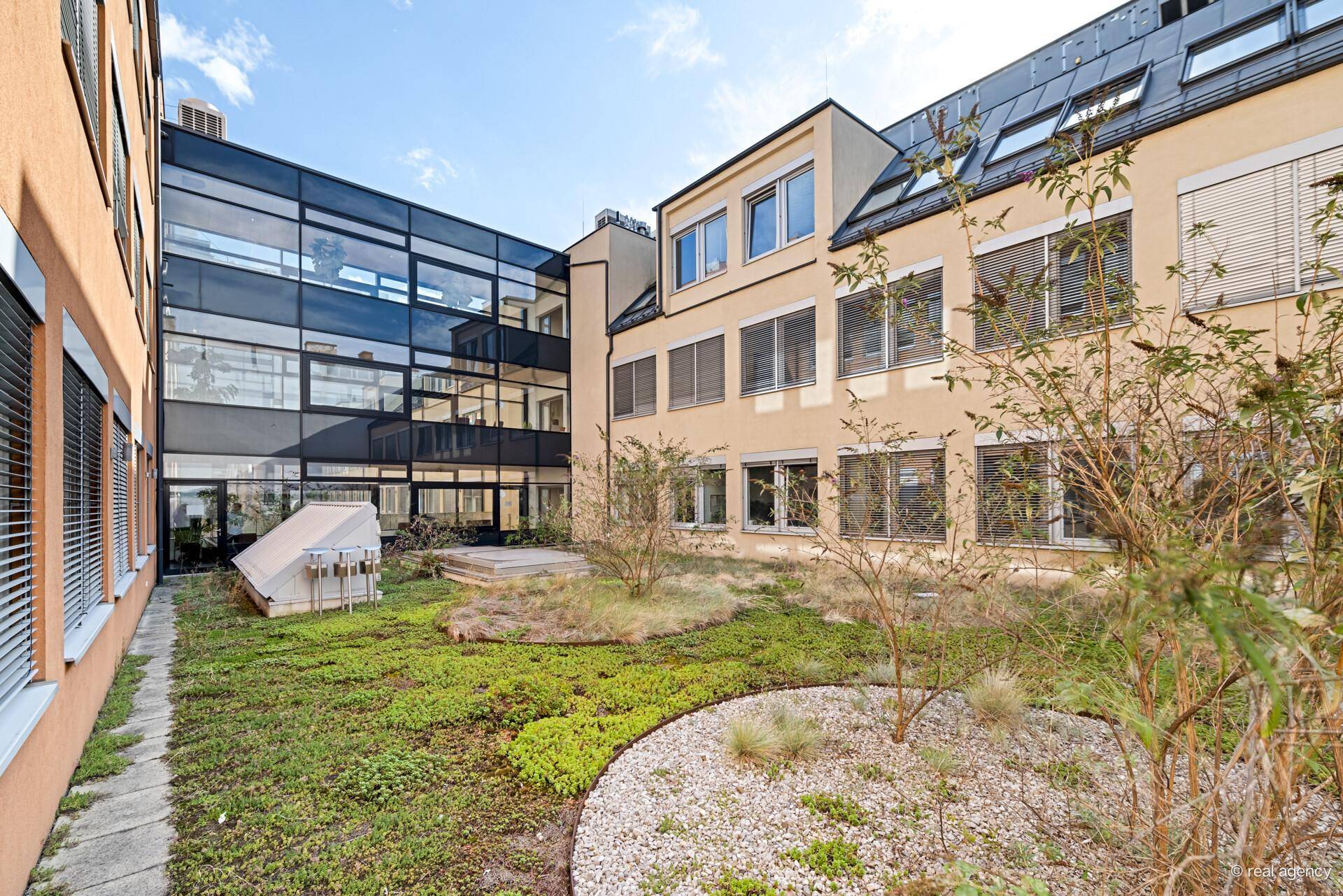 Begrünter Innenhof mit verschiedenen Pflanzen und einem modernen Glasgebäude im Hintergrund.