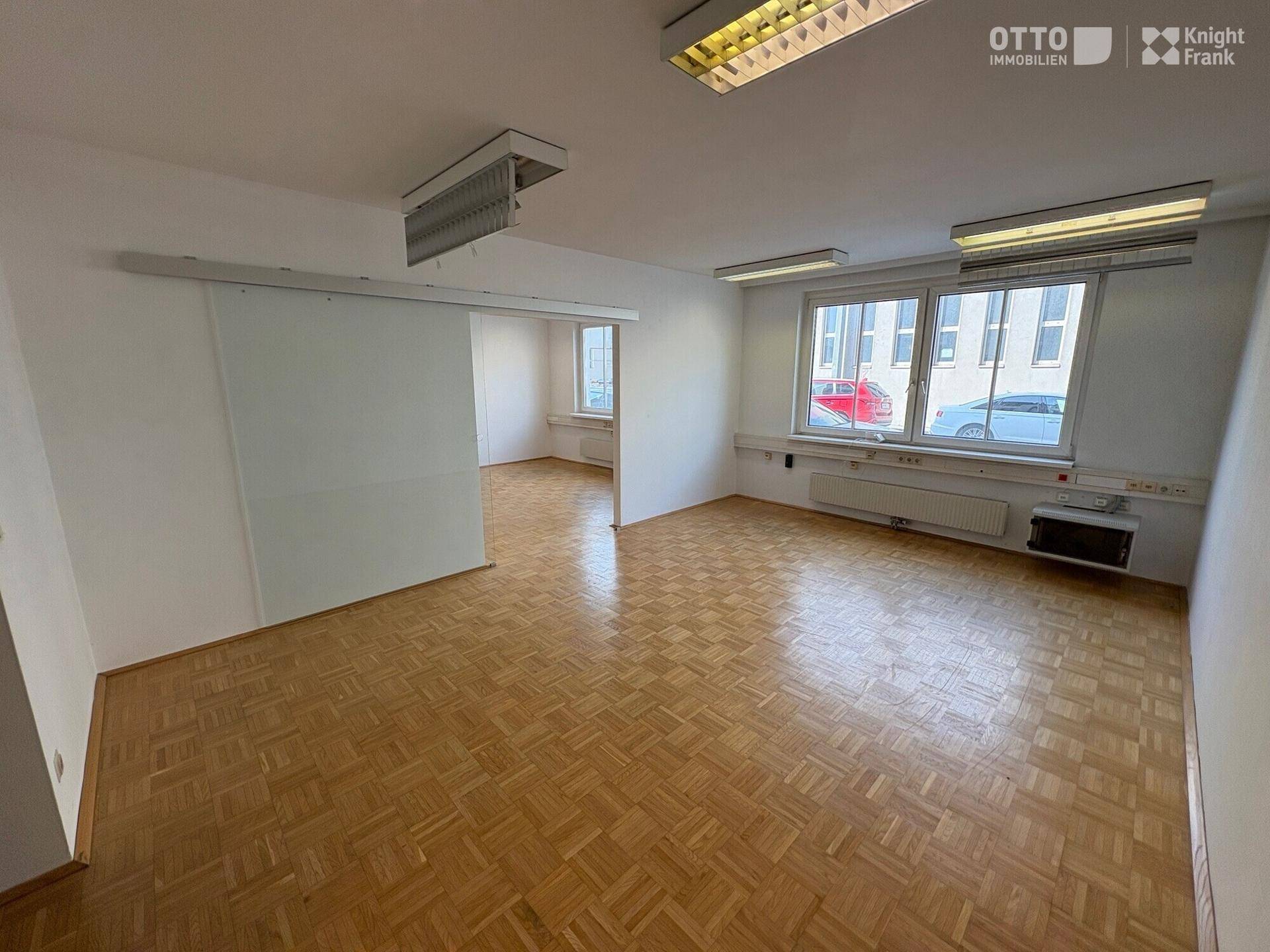 Geräumiger Bürobereich mit Parkettboden, Schiebetür und Blick auf die Straße durch große Fenster.