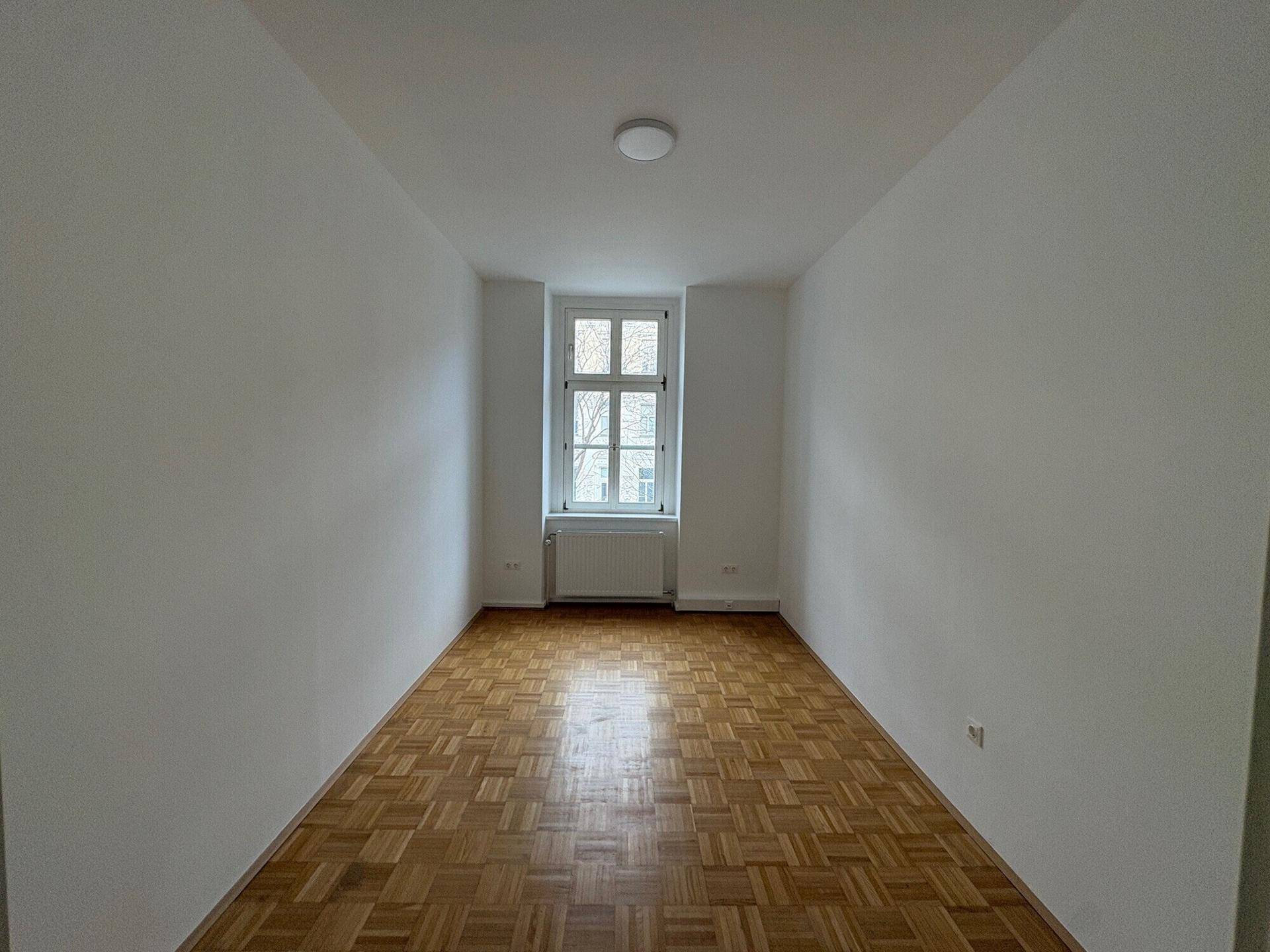 Heller Büroraum mit Parkettboden und einem einzelnen Fenster, das den Raum gut belichtet.